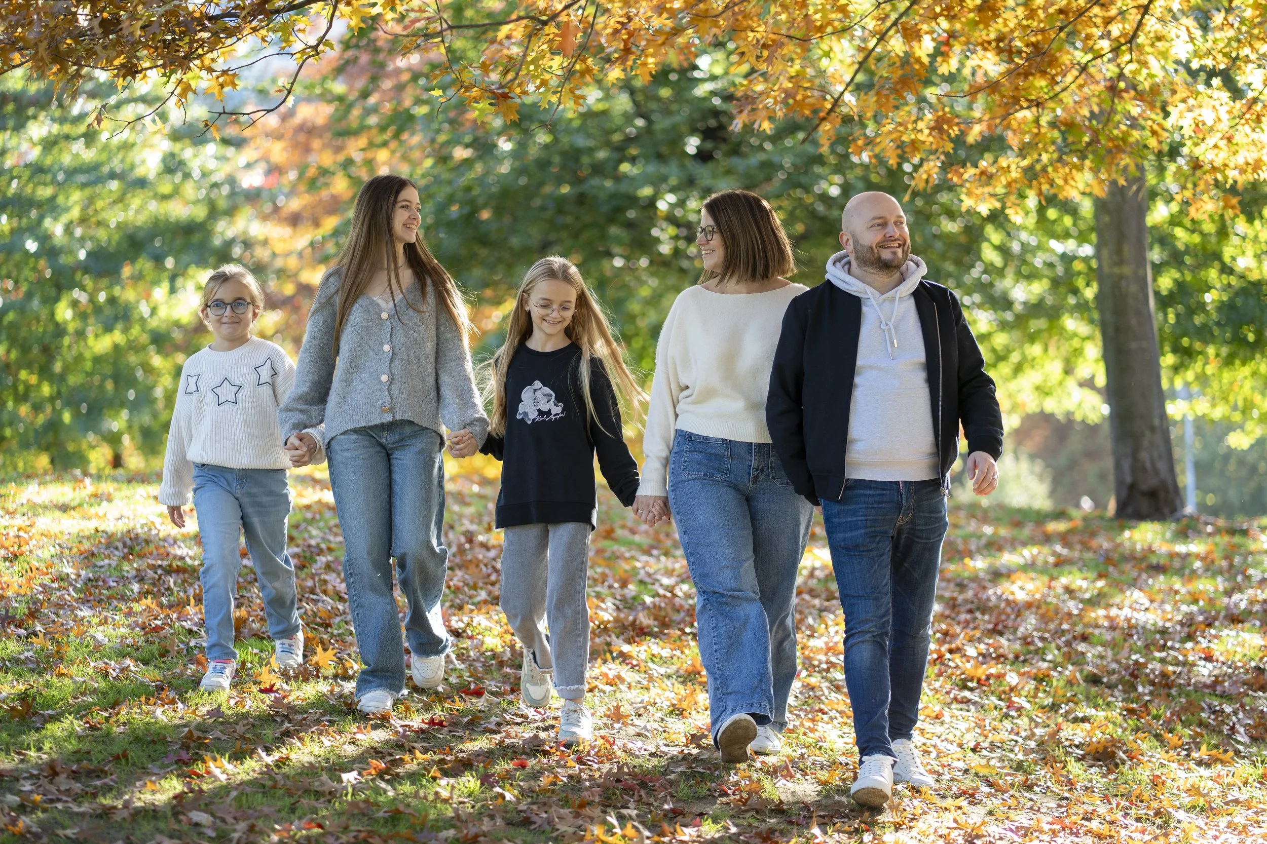 Family walking in a park during autumn, with colorful fall leaves on the ground and trees with orange and green foliage.