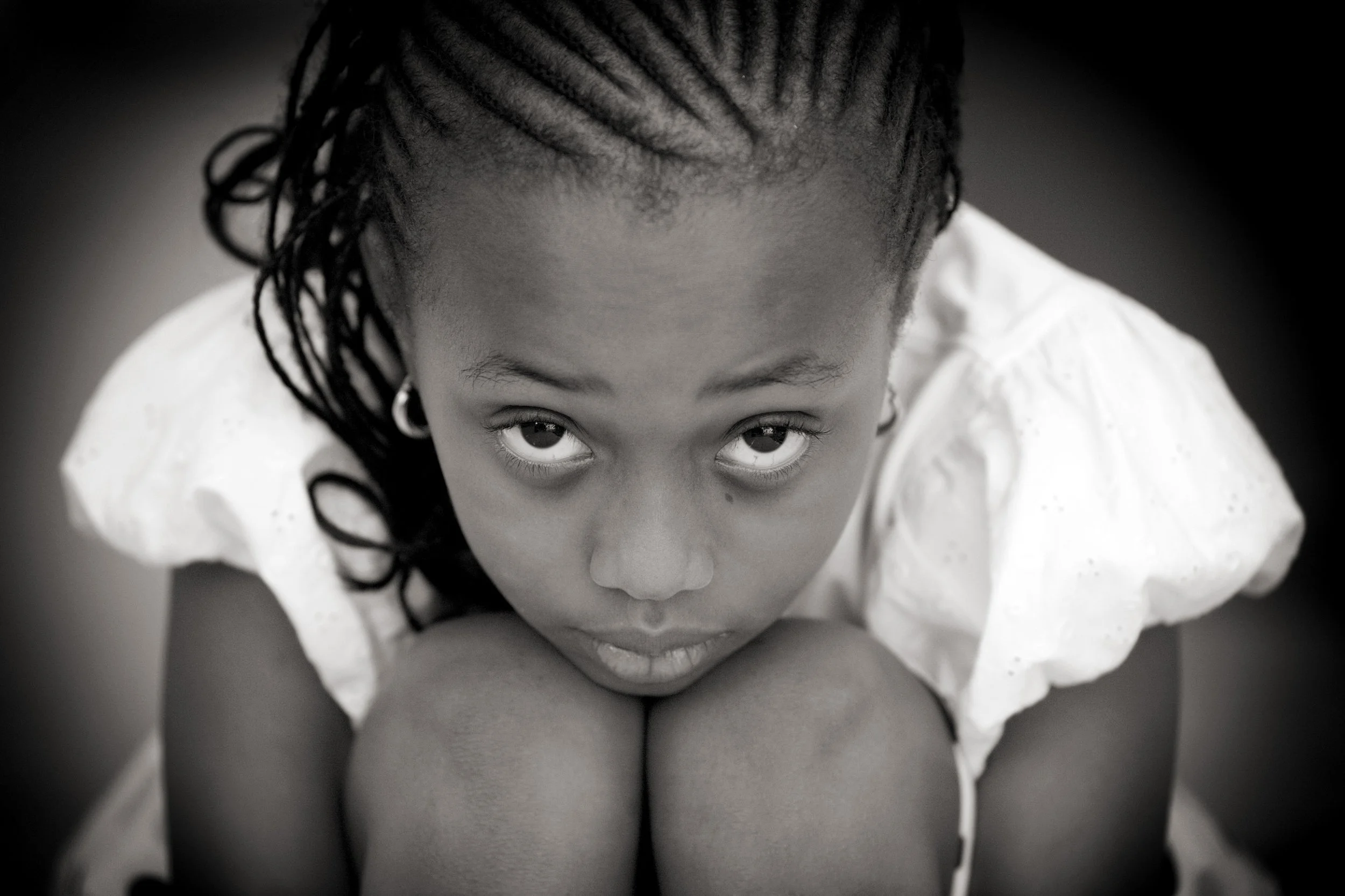 A young girl with braided hair looking up at the camera, sitting with her knees close to her chest, wearing a white top.