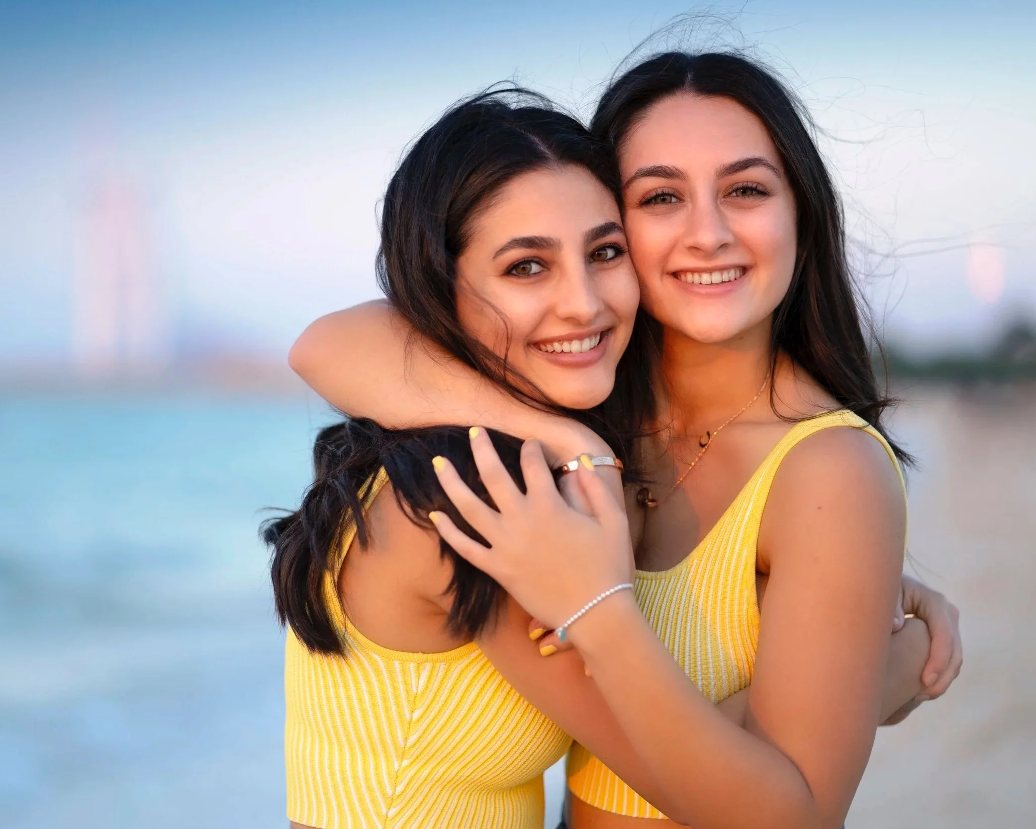 Two young women with dark hair smiling and hugging at the beach during sunset, wearing matching yellow tops.