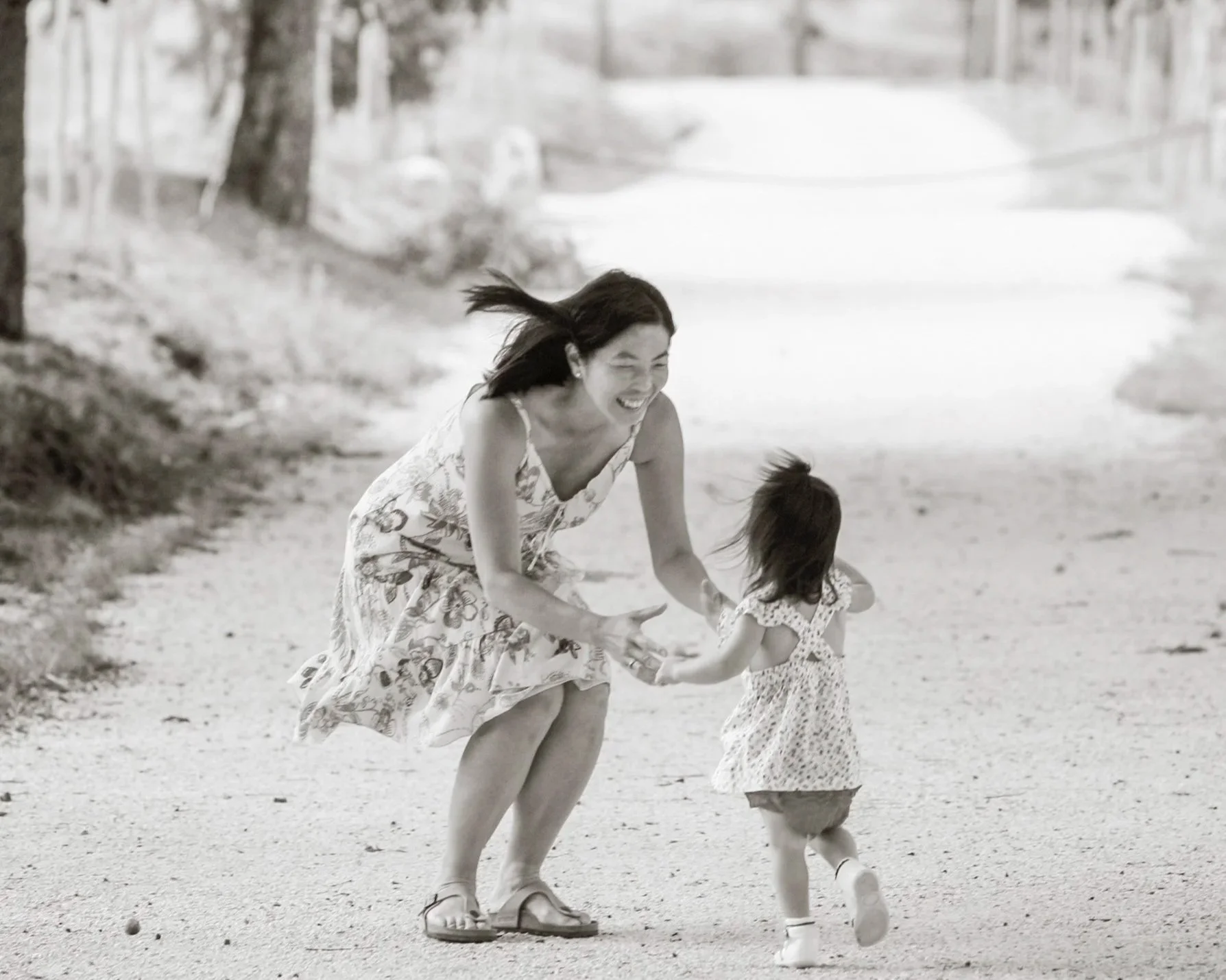 A woman in a floral dress joyfully playing with a young girl in a park or outdoor setting, both holding hands and running.