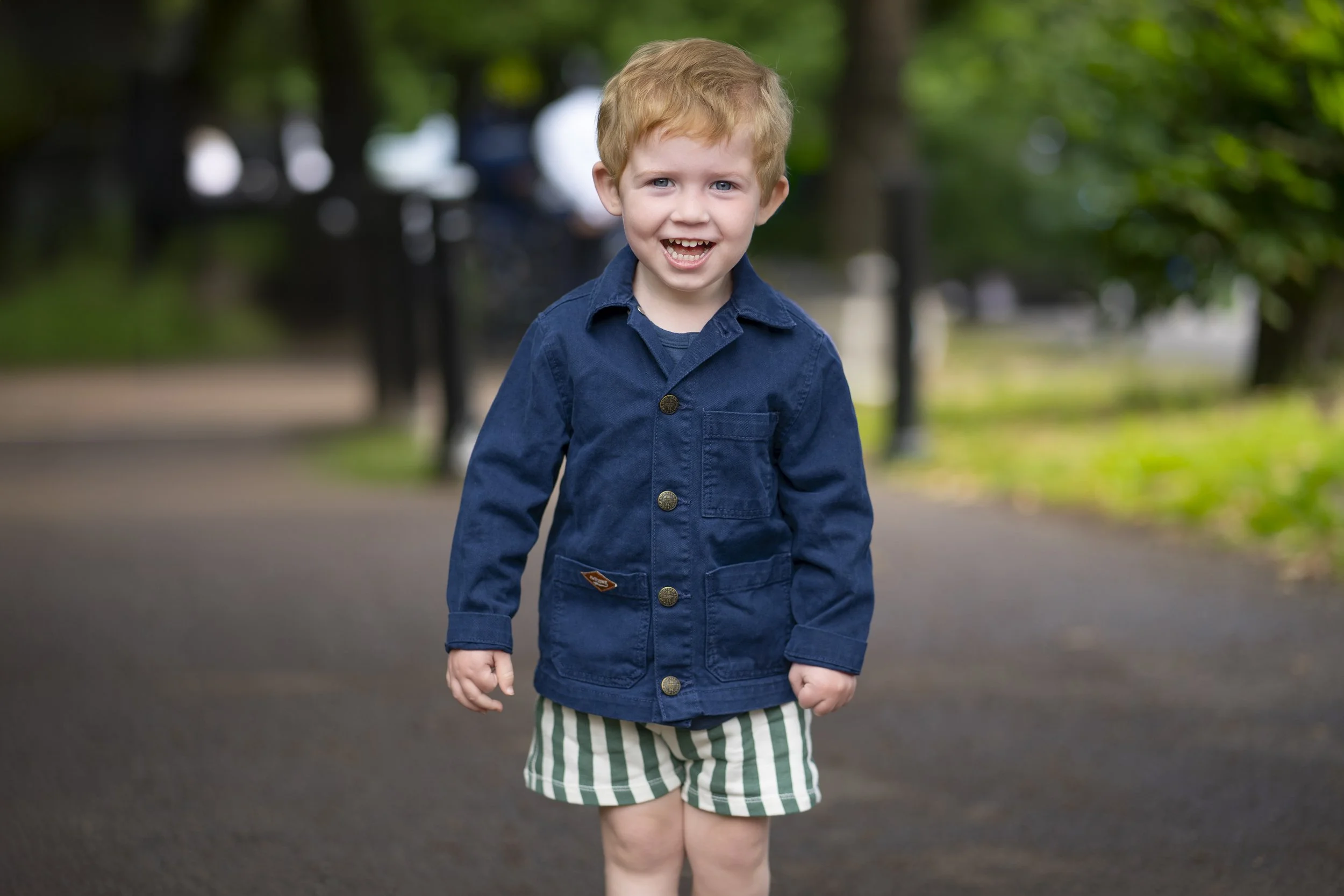 A young boy with red hair smiling and walking outdoors on a paved path with green trees in the background, wearing a blue jacket and green-and-white striped shorts.
