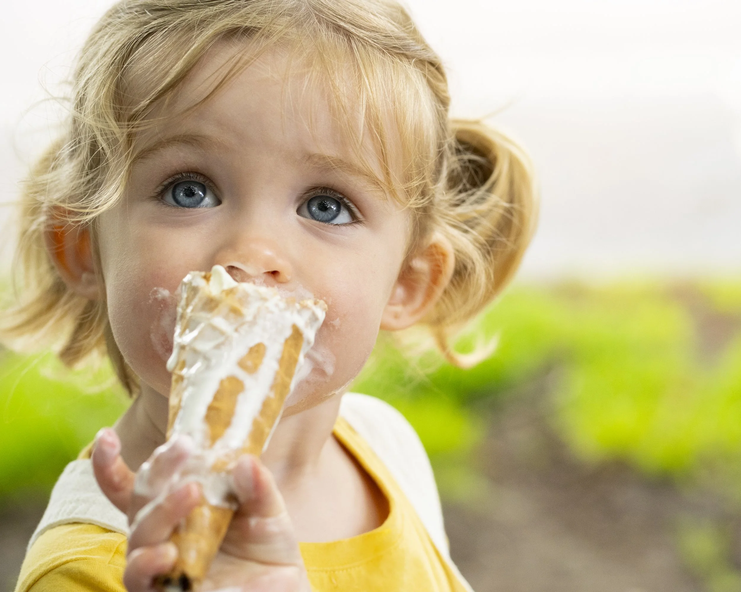 A young girl with blonde hair and blue eyes eating a partially eaten ice cream cone with vanilla ice cream and caramel topping outdoor with green foliage in the background.