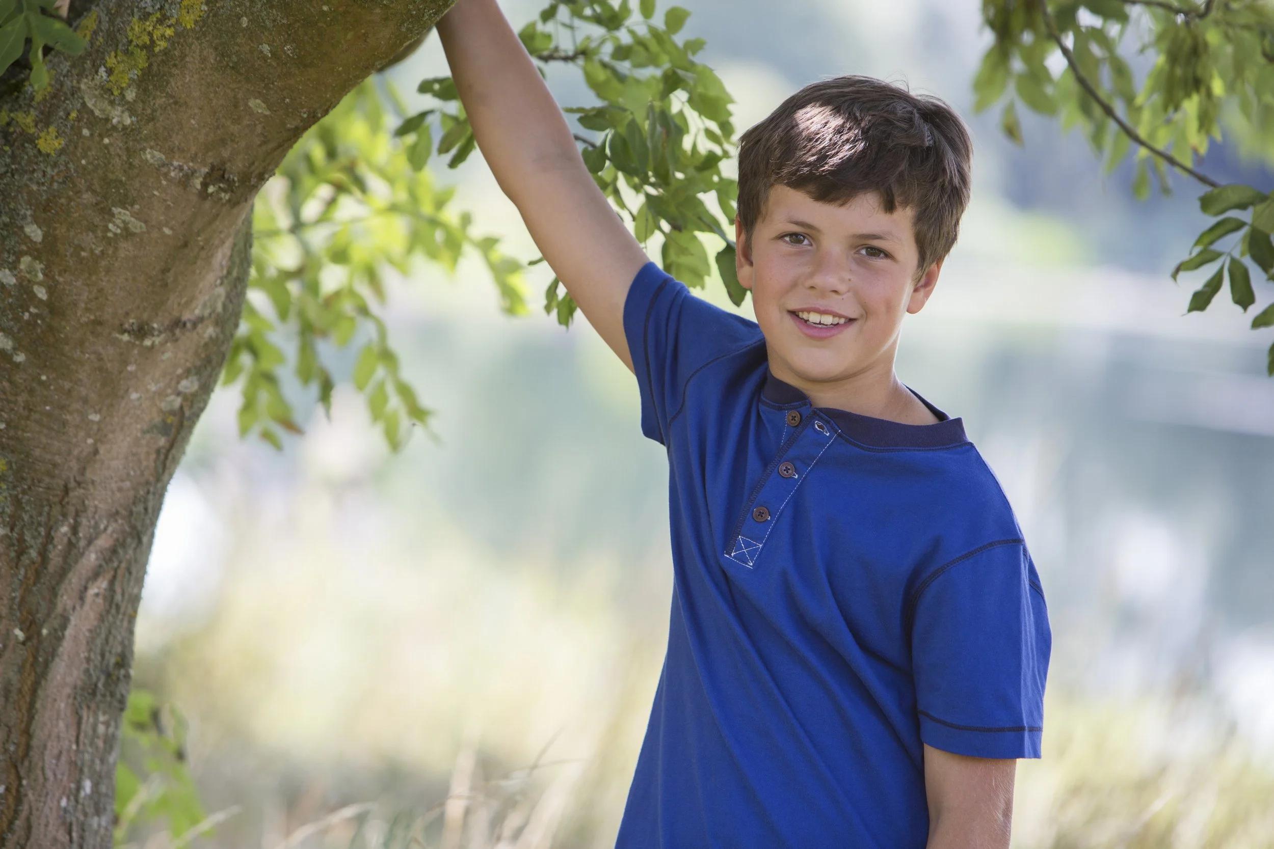 A young boy with short brown hair wearing a blue shirt, smiling, standing next to a tree near a body of water with green leaves and a blurred background.