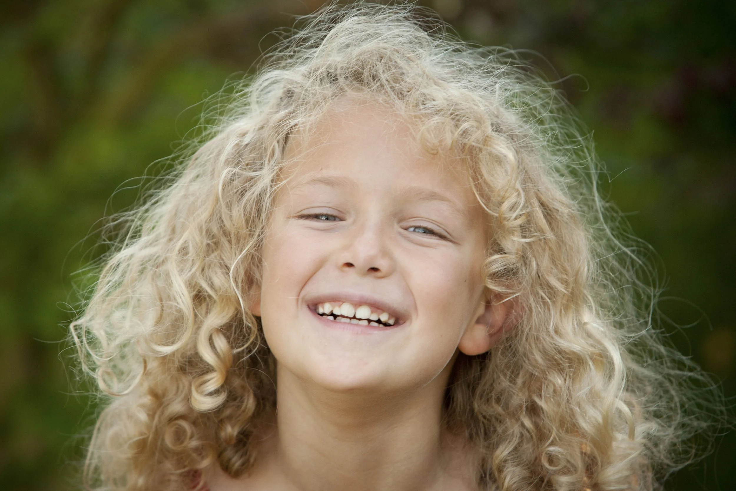 A young girl with curly blonde hair smiling outdoors against a blurred green background.