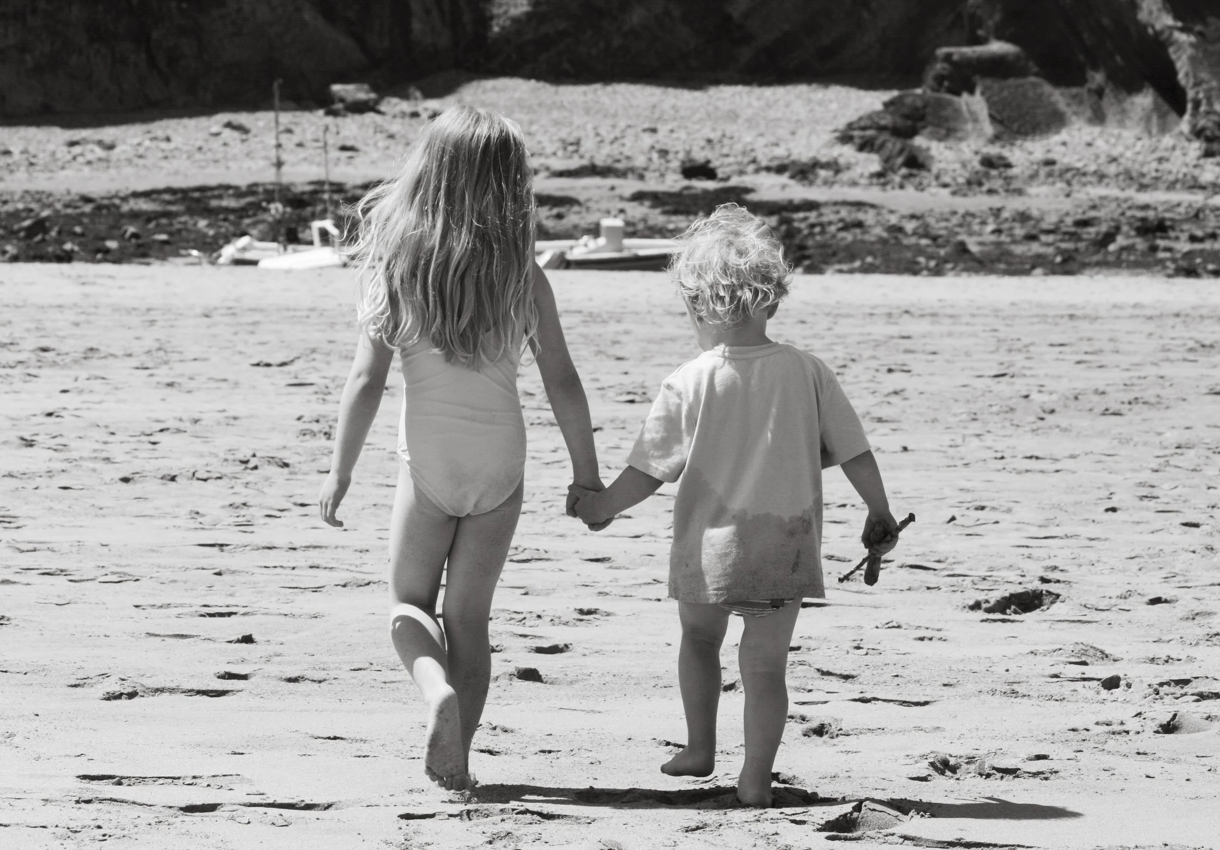 Two young children, a girl and a boy, holding hands and walking barefoot on a sandy beach toward the water.