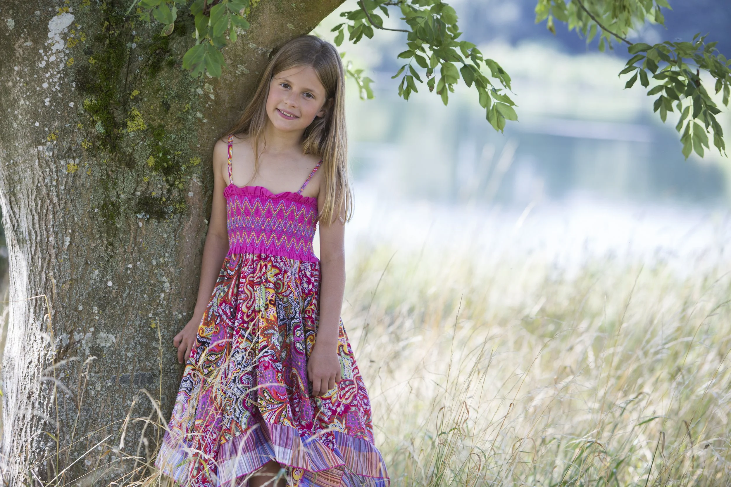 A young girl with long blonde hair wearing a colorful pink and patterned dress, standing next to a tree in a grassy outdoor setting with a lake in the background.