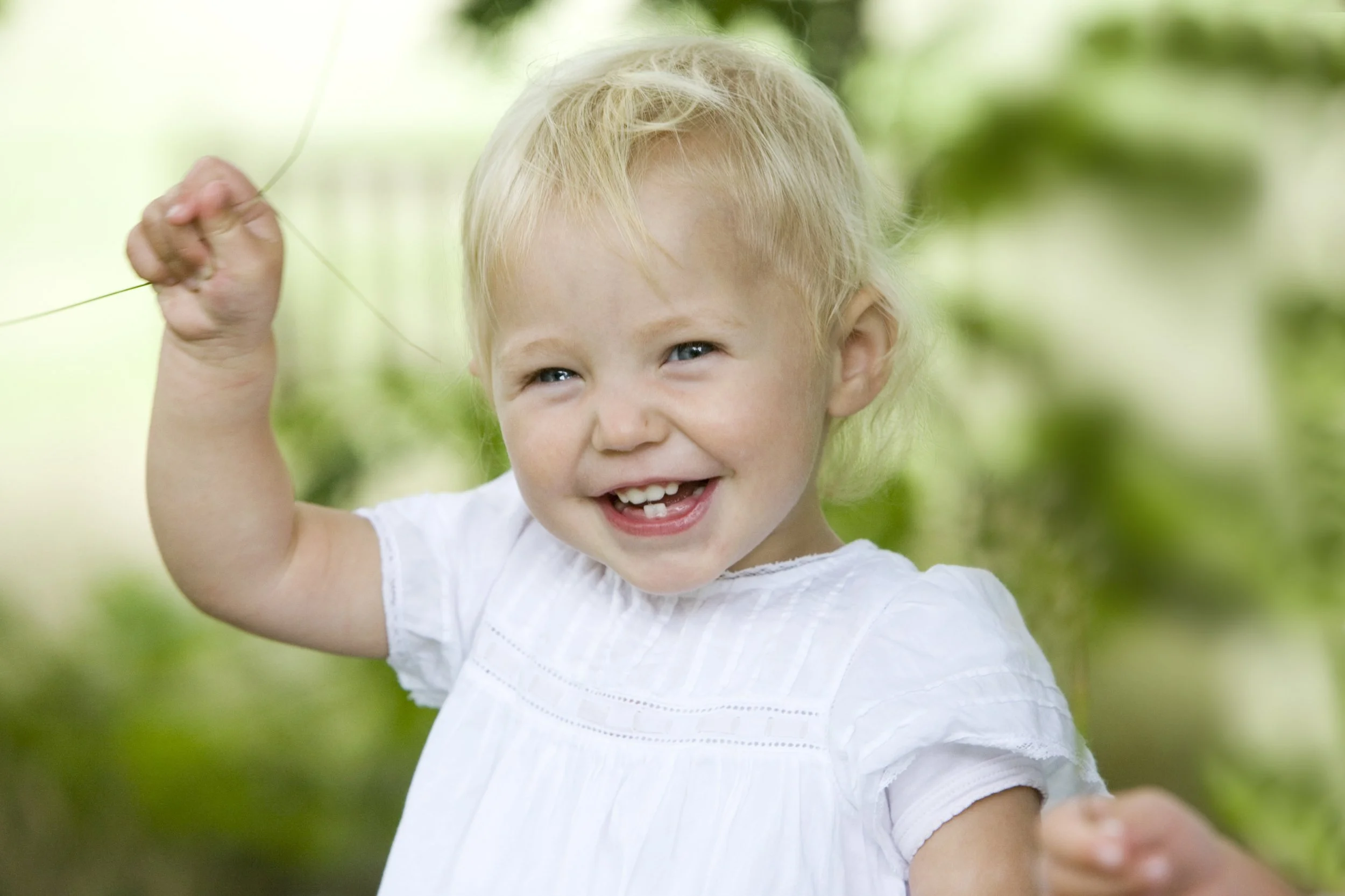 A young blonde girl smiling outdoors, holding a string or branch, wearing a white dress, with a background of green foliage.