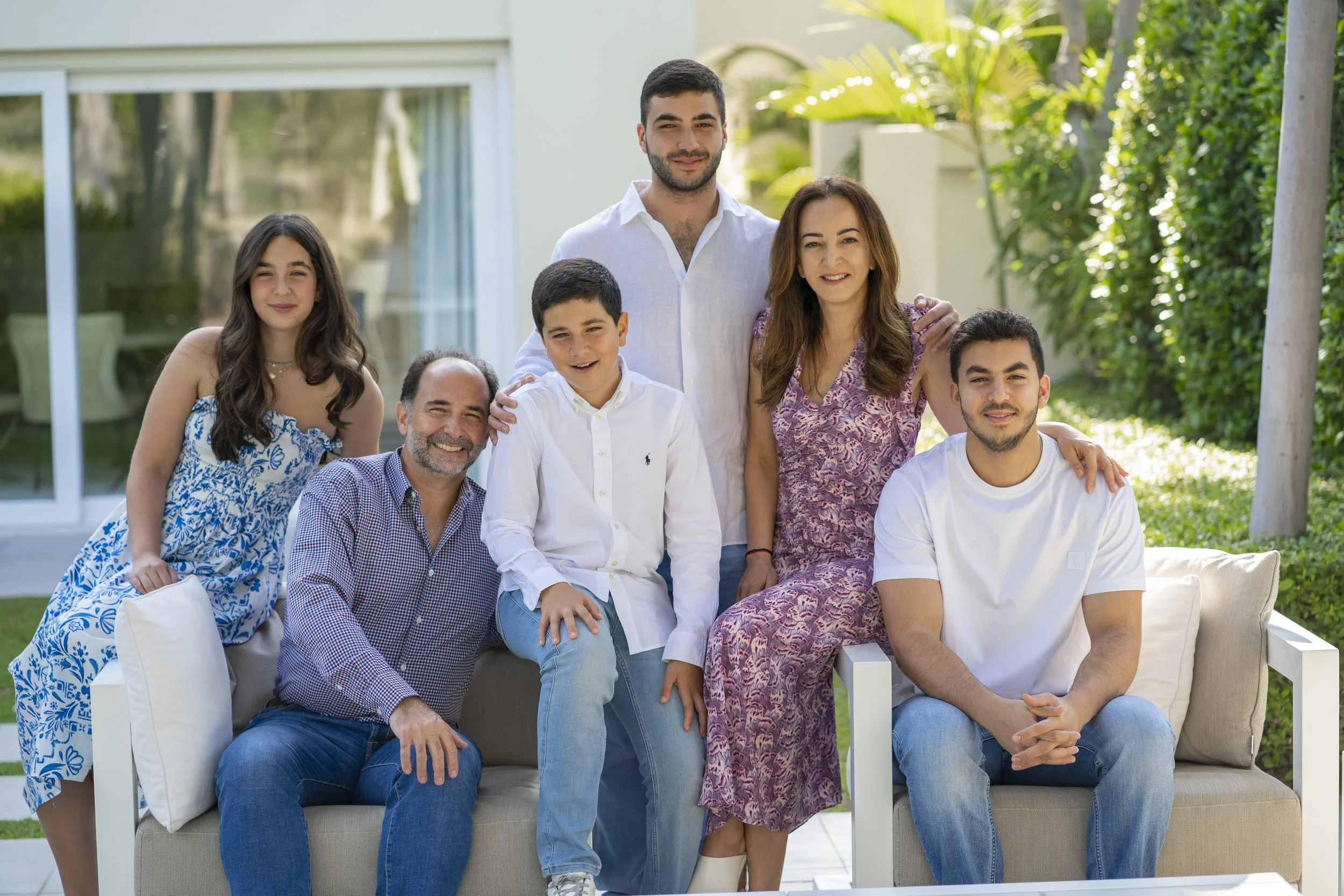 A multi-generational family of seven people posing outdoors in a garden with greenery, sitting and standing on a beige sofa and outdoors furniture. They are smiling and dressed casually for a sunny day.