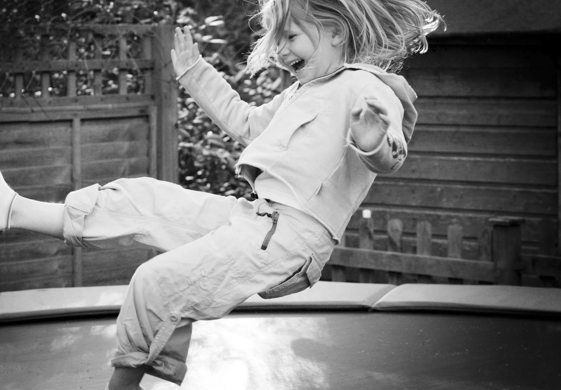 A young girl with shoulder-length hair smiling and falling backward onto a trampoline outdoors, with a wooden fence and greenery in the background.