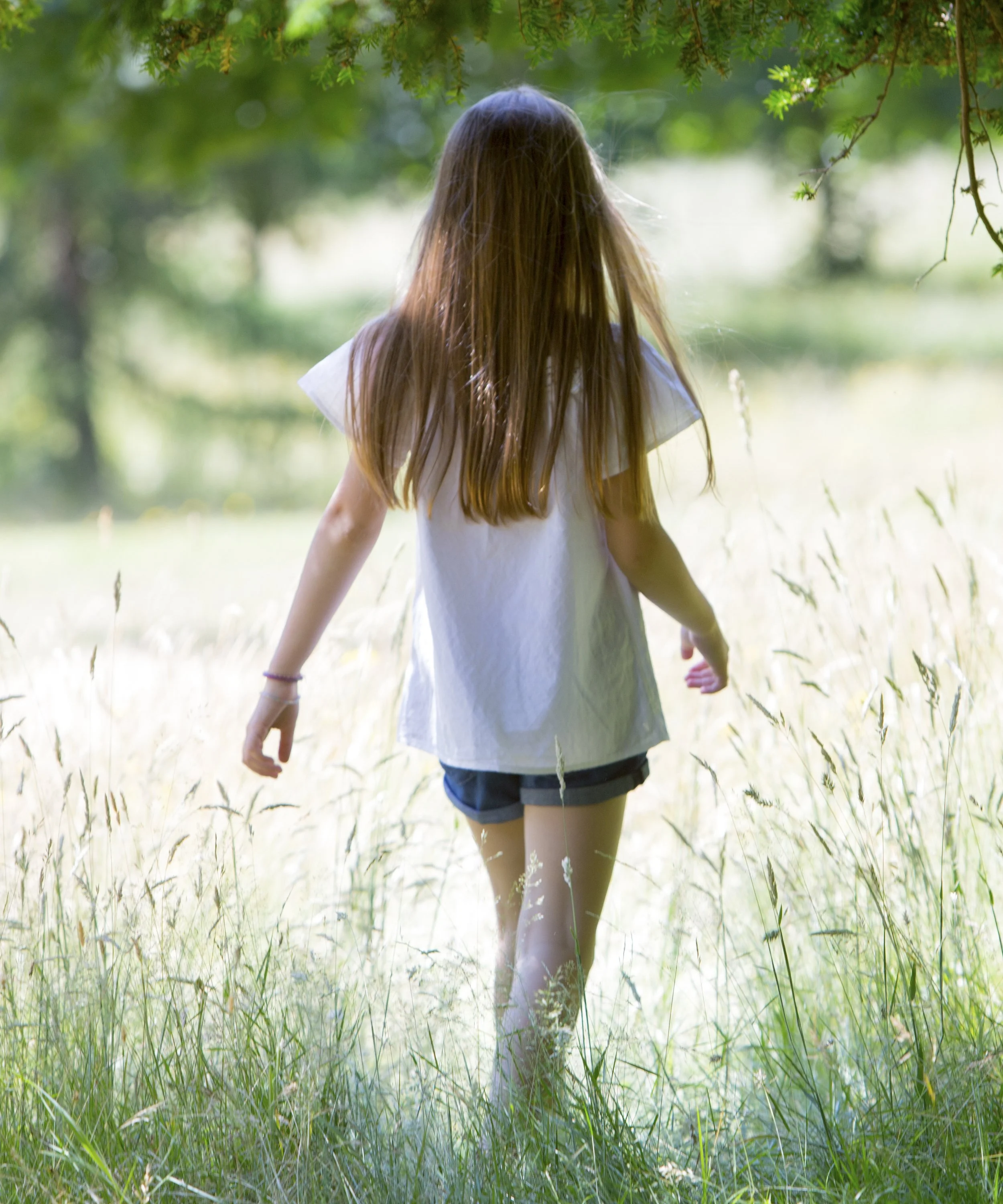 A girl with long hair in a white top and dark shorts walking through a grassy field near trees.