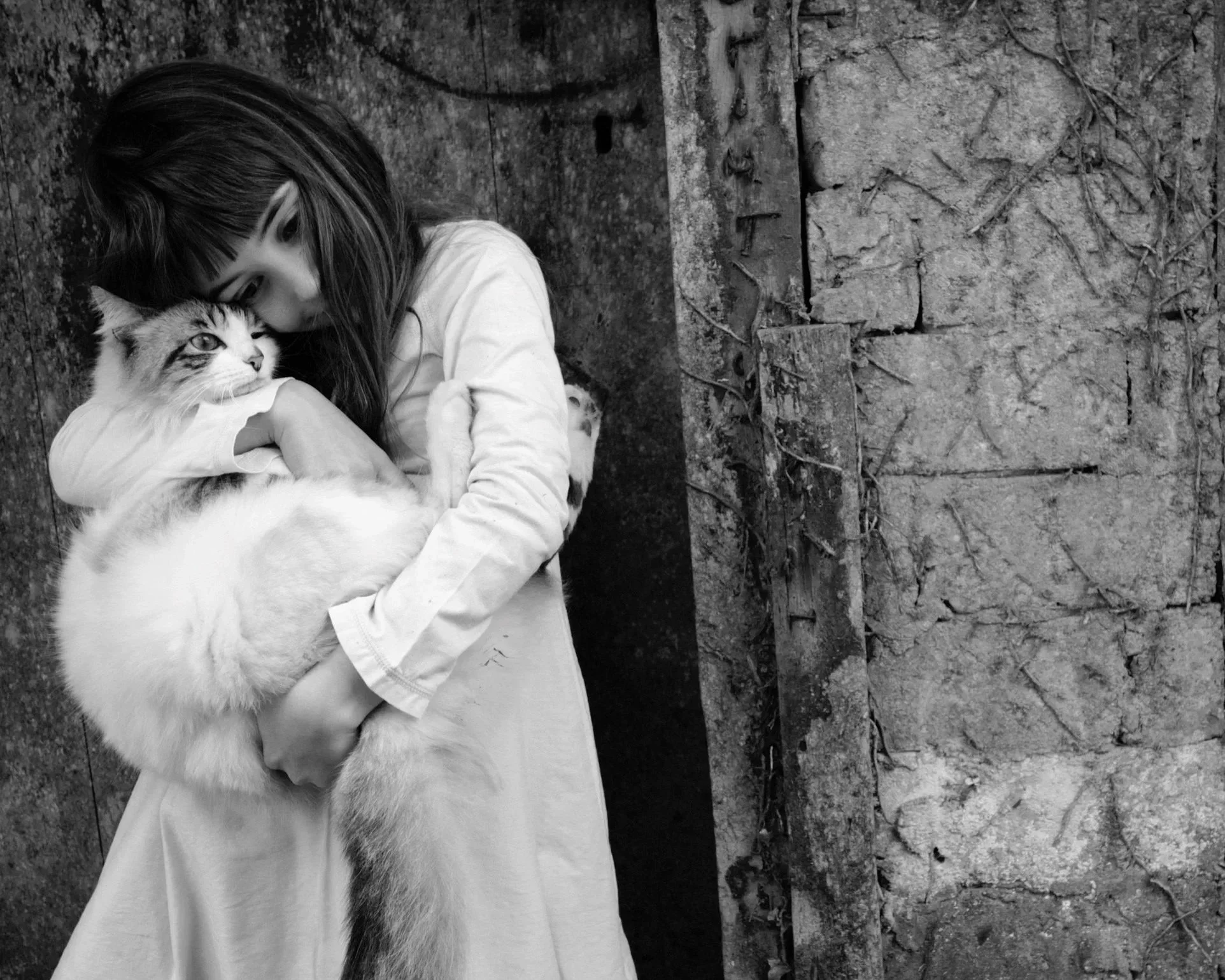 A young girl with long dark hair holding a fluffy cat while standing against an old textured stone wall, in black and white.