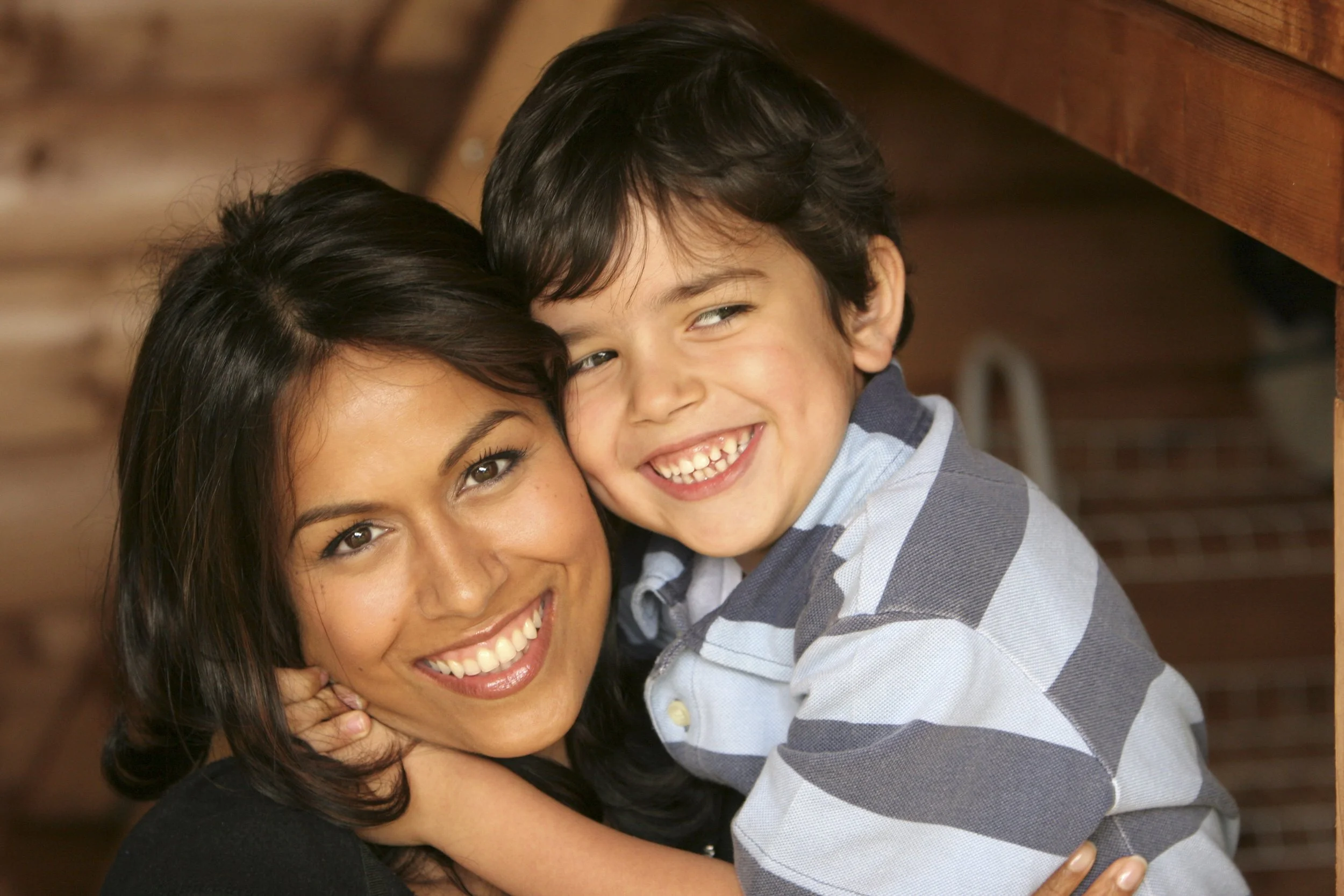 A woman and a young boy are hugging each other, smiling happily indoors with a wooden background.