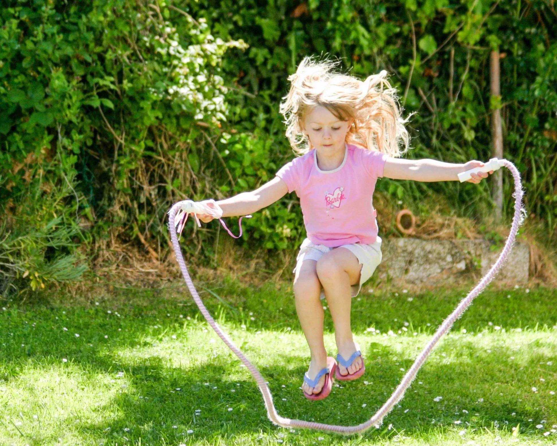 A young girl jumping on a pink jump rope outdoors on a grassy lawn with green bushes in the background.