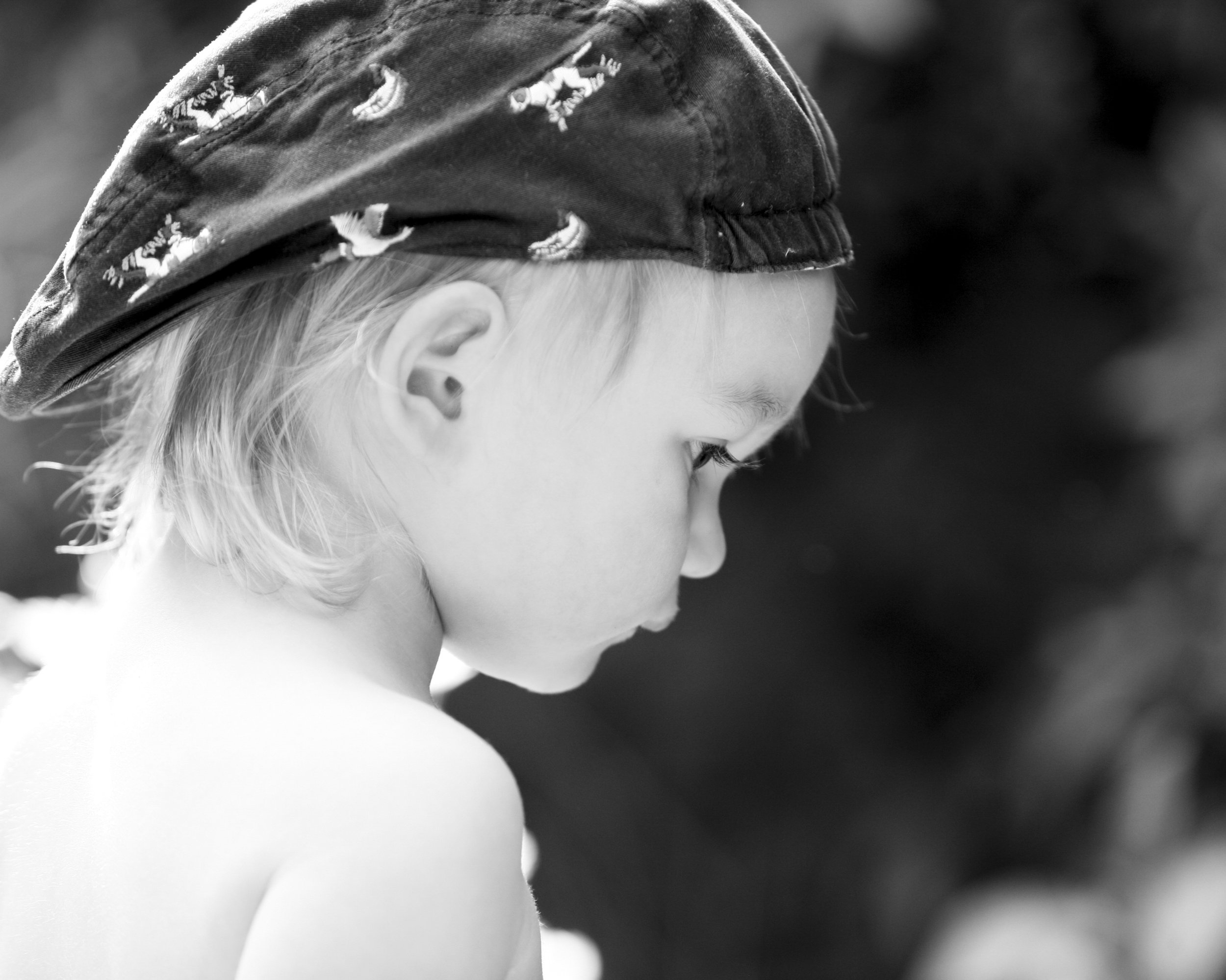 A young child with light hair wearing a patterned cap, looking downward with a serious or contemplative expression, in black and white.