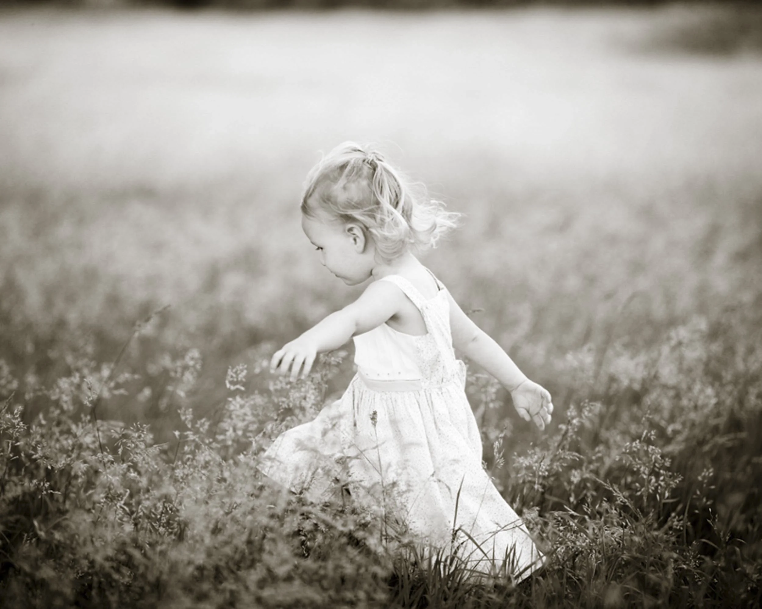A young girl with curly hair in a sleeveless dress walking through a grassy field in black and white.