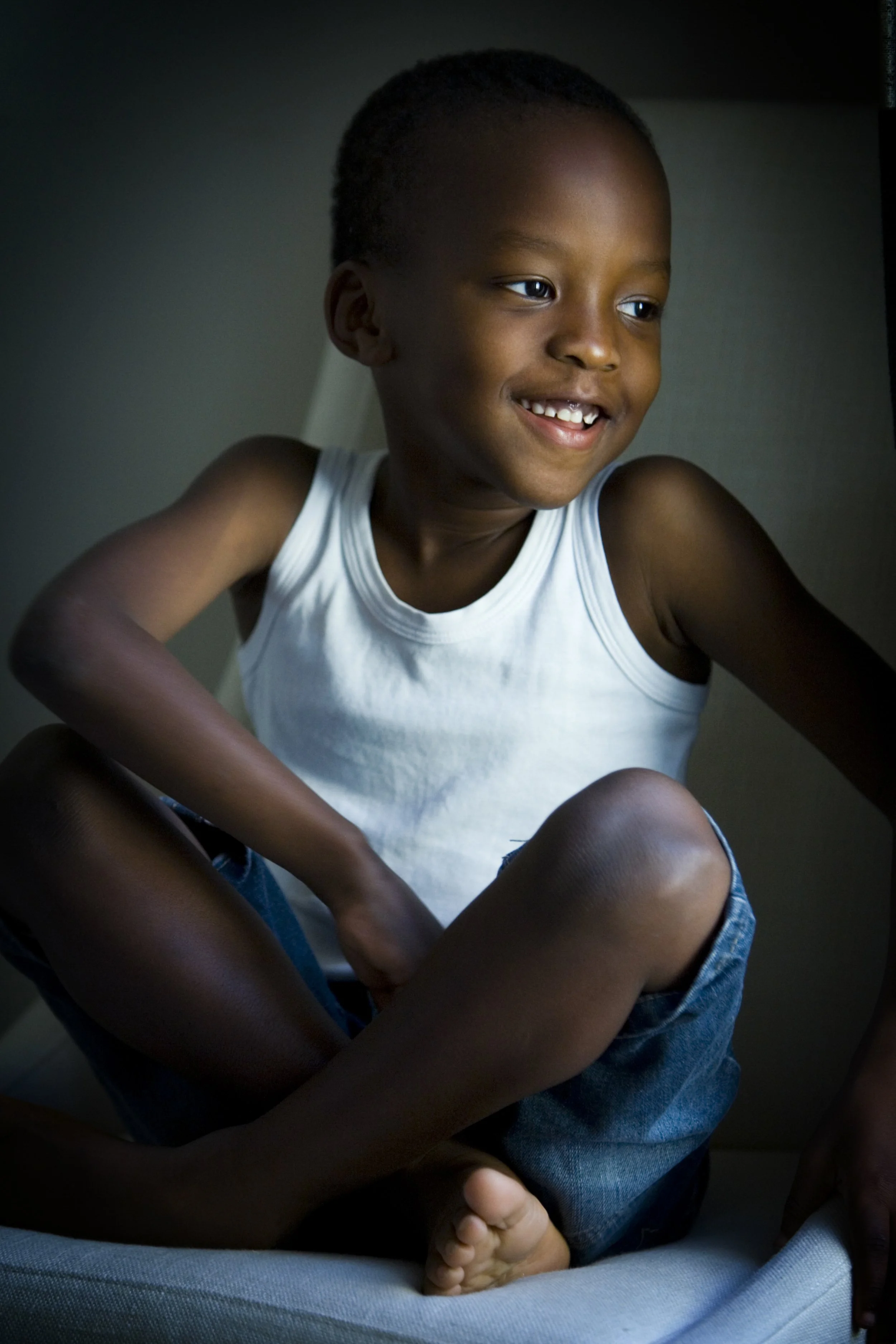 A young boy with dark skin and short hair, smiling and sitting cross-legged on a chair, wearing a white tank top and denim shorts, with a dark and softly lit background.