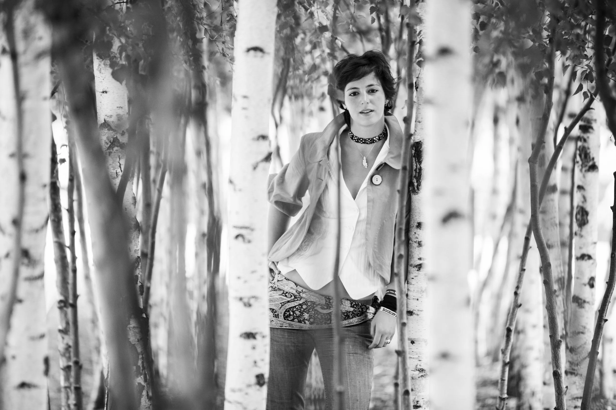 A woman with short dark hair leaning against a tree in a forest of birch trees.