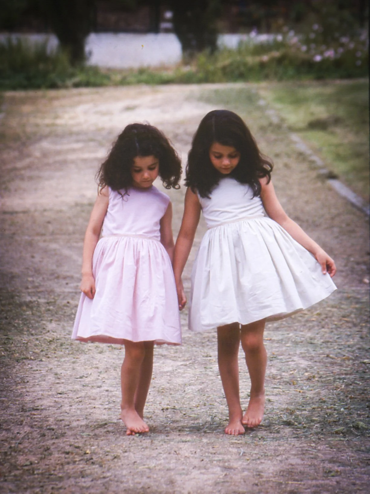 Two young girls walking barefoot on a dirt path, holding hands, wearing light-colored dresses, and looking down.