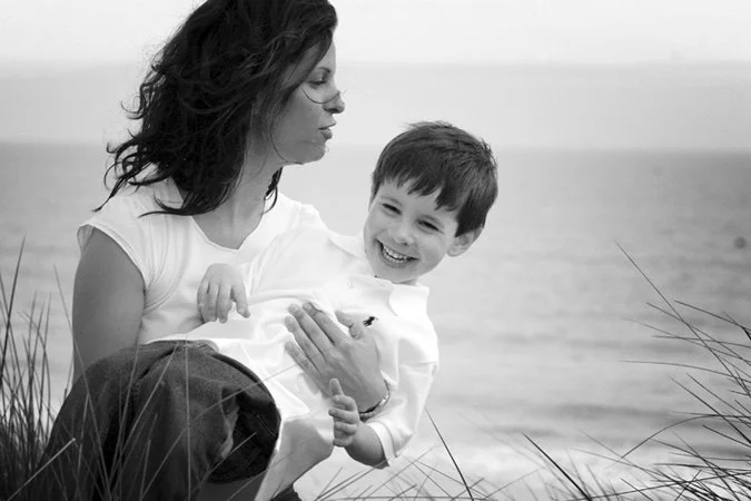 A woman holding a smiling young boy by the beach, sitting in tall grass.