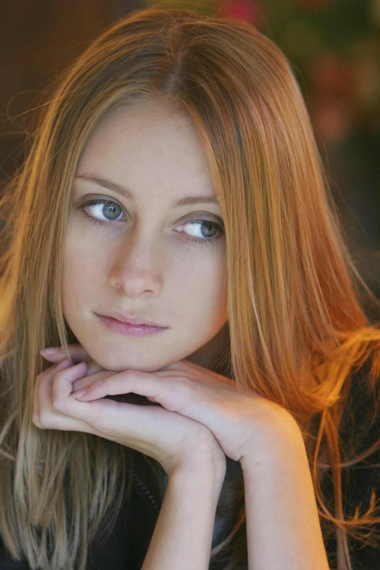 Close-up portrait of a young woman with long red hair and blue eyes resting her chin on her hands, looking to the side with a soft expression.