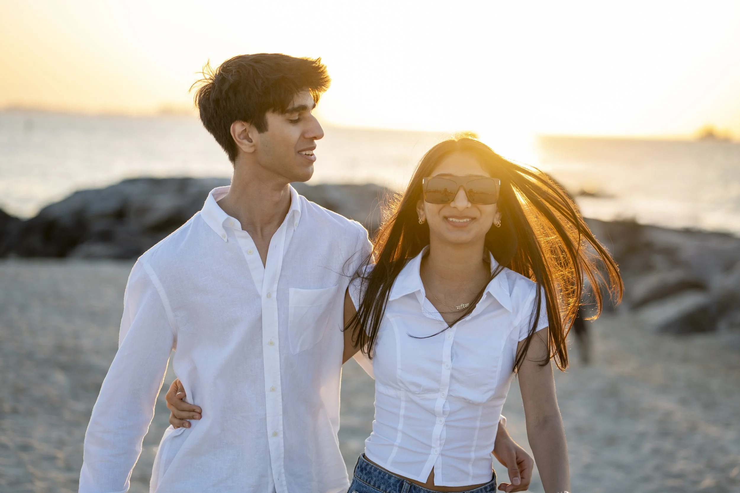 A young man and woman walk together on a beach during sunset, smiling. The man has dark hair and wears a white shirt, while the woman has long brown hair, wears sunglasses, and a white shirt.