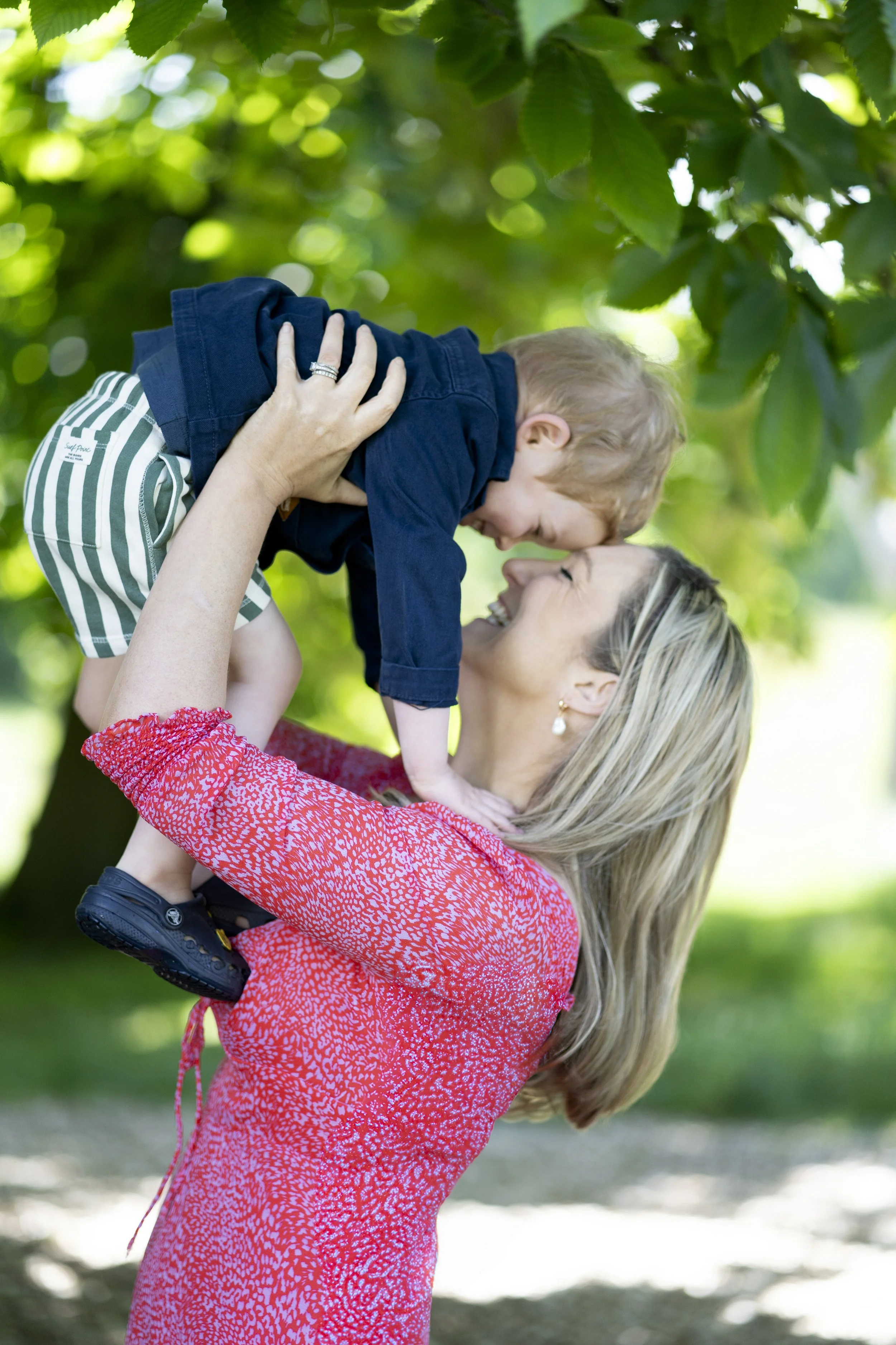 A woman lifting a young boy in the air outdoors, both smiling and touching foreheads amidst green trees.