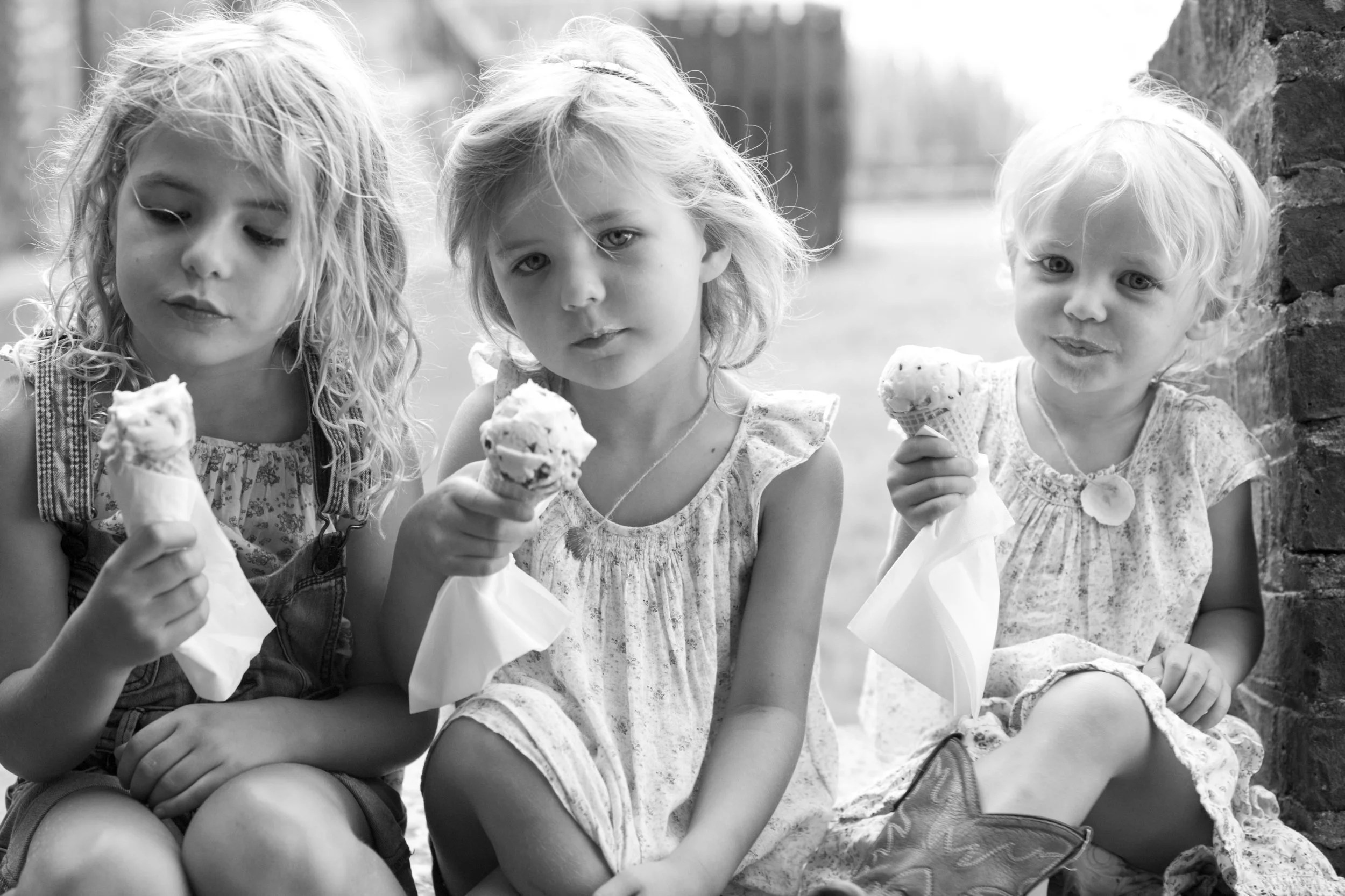 Three young girls sitting outdoors, each holding ice cream cones, with a brick wall and blurred background.