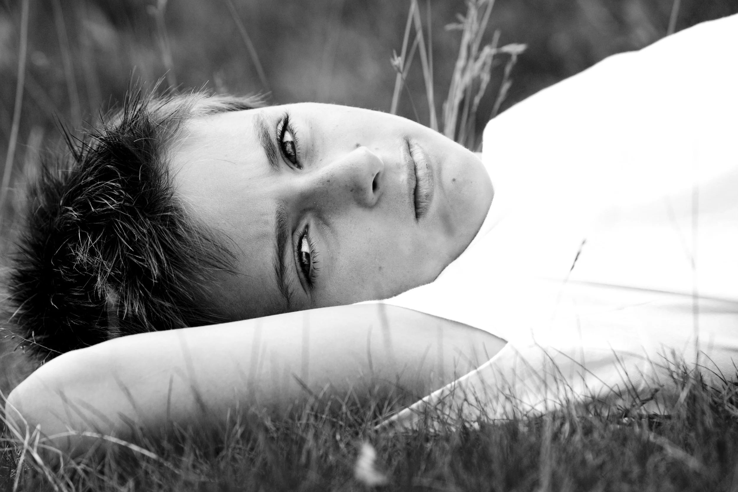 Black and white photo of a woman lying on the grass, with her head resting on an arm, gazing at the camera.
