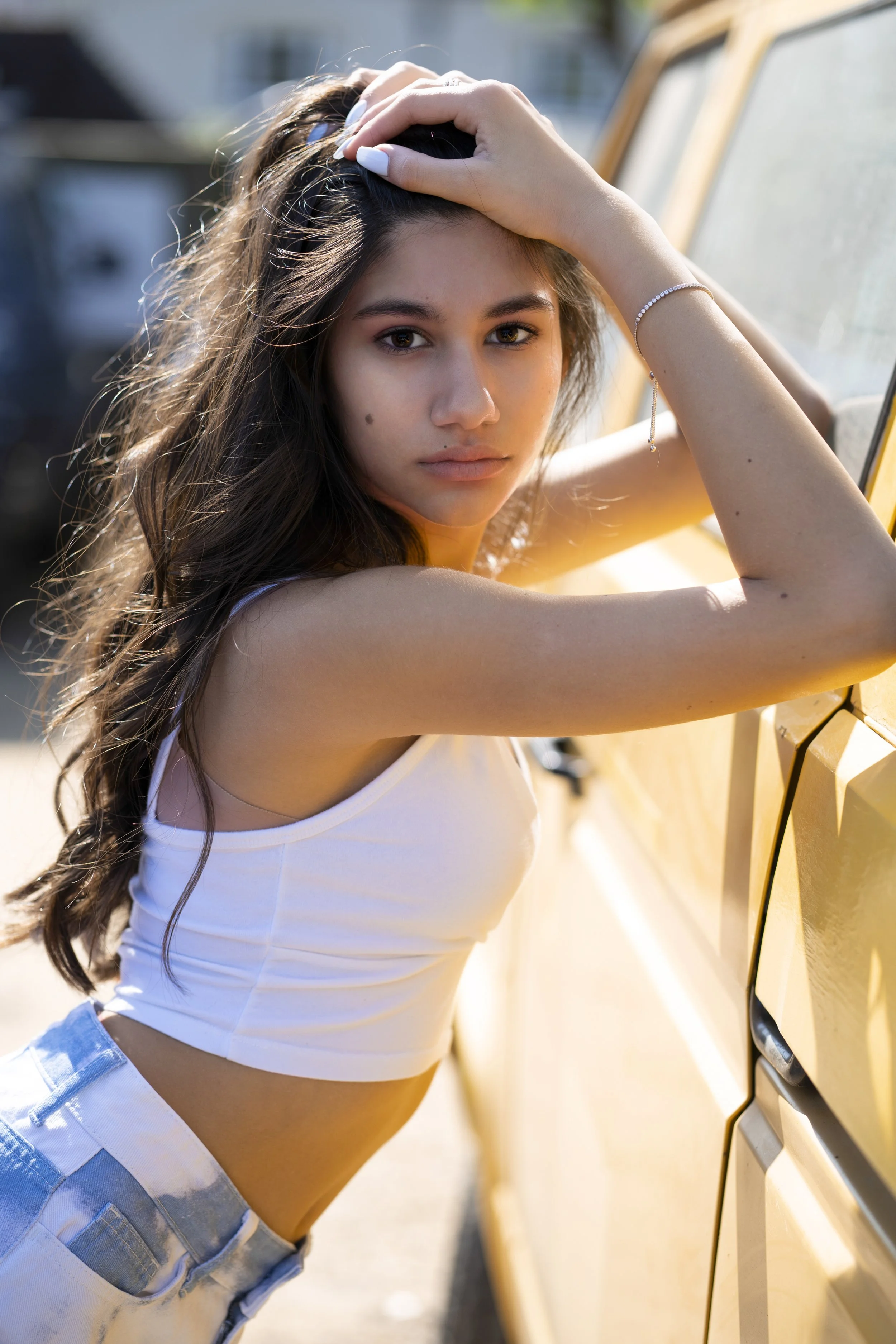 A young woman with long wavy hair, wearing a white sleeveless crop top and light blue jeans, leans against a yellow vehicle with her hand on her head, looking into the camera with sunlight highlighting her face.