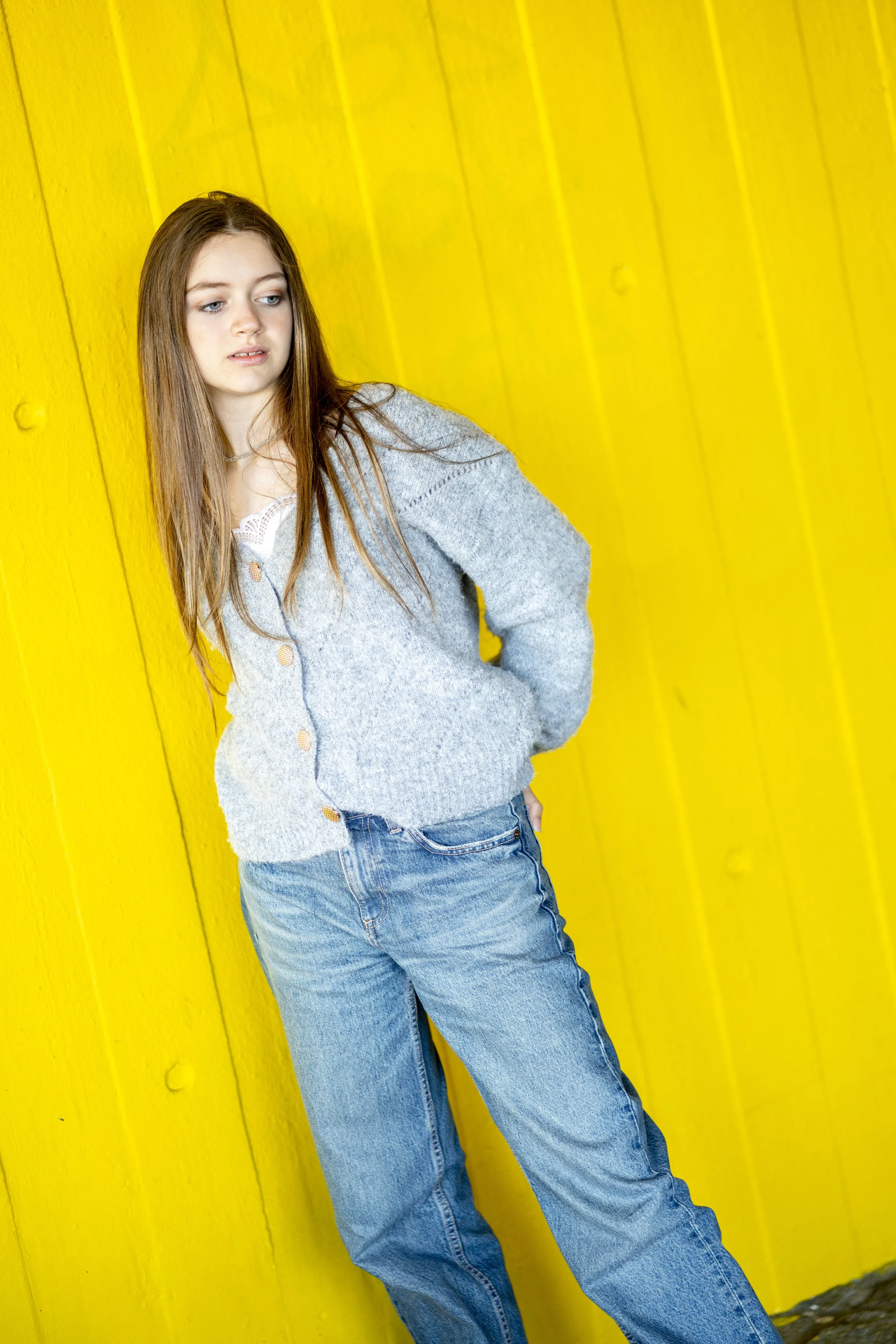 A young woman with long brown hair wearing a gray sweater and blue jeans, standing against a bright yellow wooden wall.