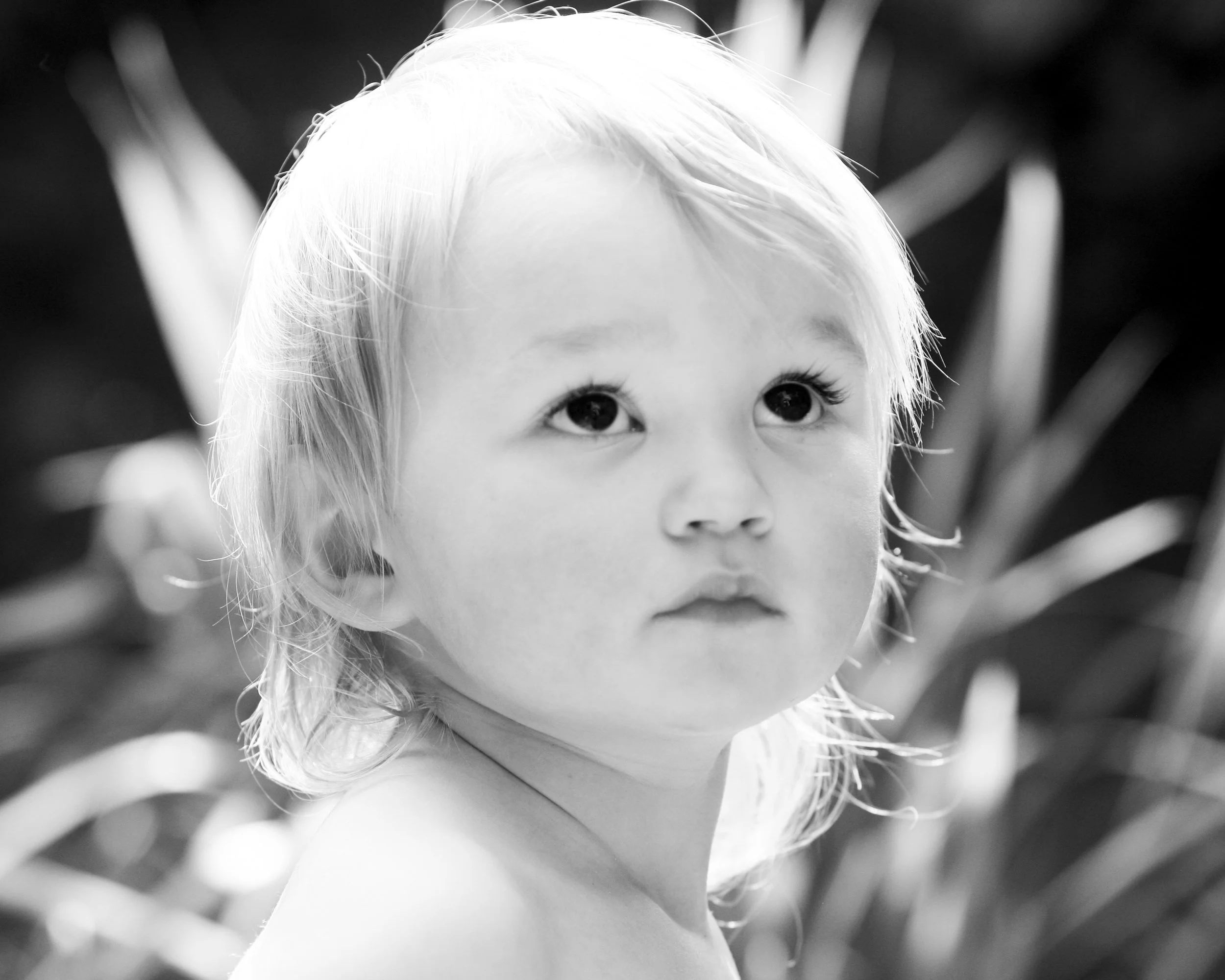 A young child with light hair and large eyes looking upwards, with blurred plants in the background.