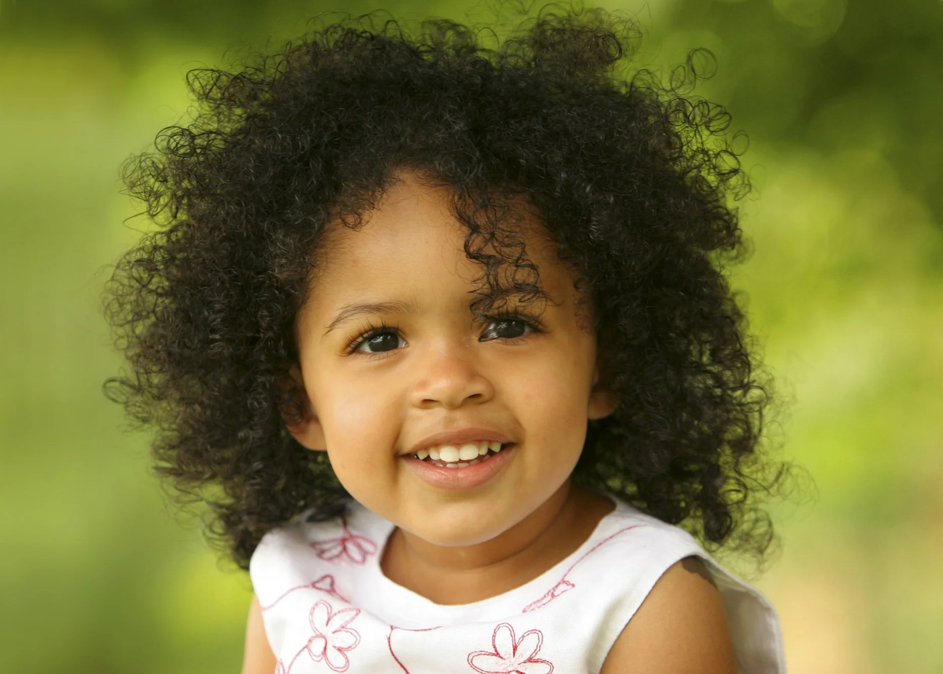 A young girl with curly dark hair smiling outdoors with a blurred green background.