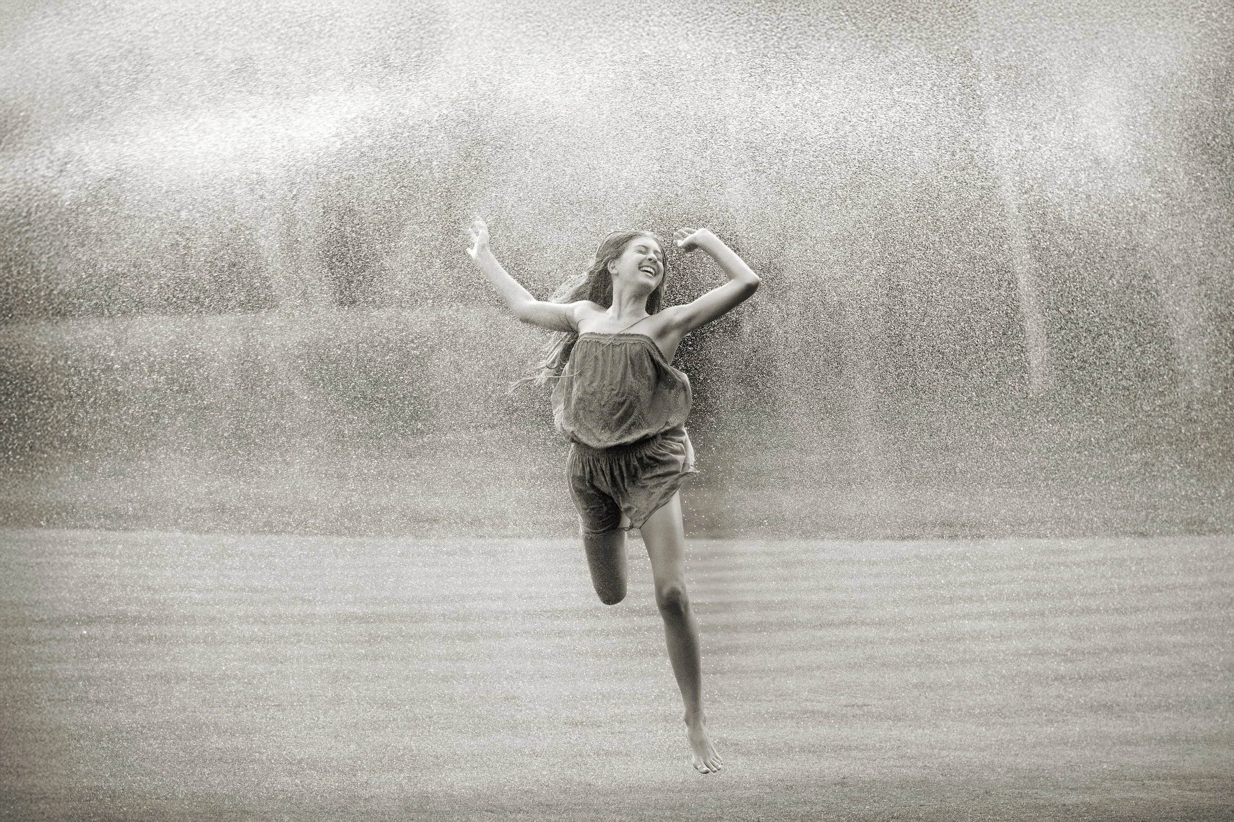 A woman is joyfully jumping in water with a big splash around her, smiling and with arms raised.