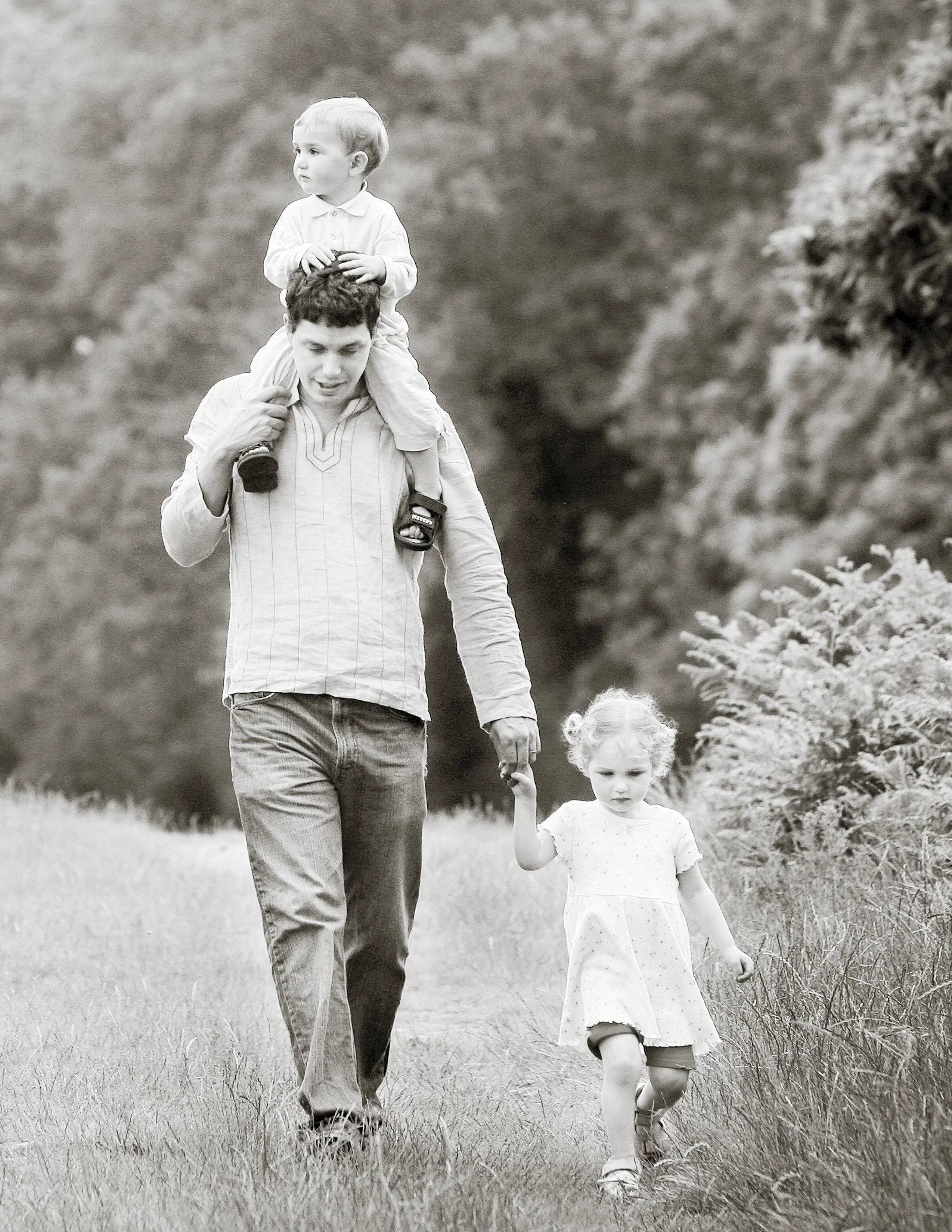 A man walking outdoors with two young children, one on his shoulders and holding his hand, in a natural setting with trees in the background.