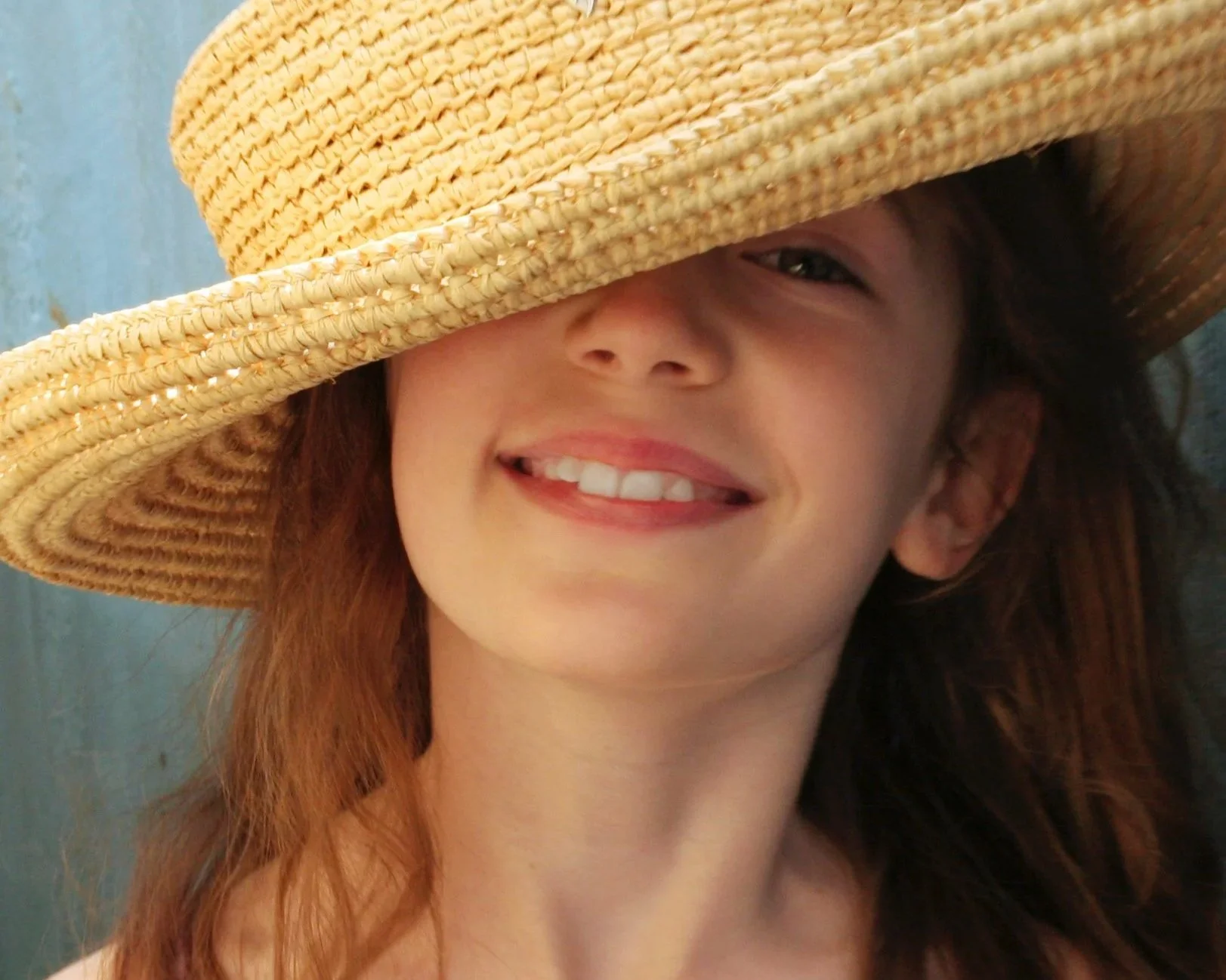A woman with long brown hair and a bright smile, wearing a large straw hat that partially covers her face, standing outdoors against a blue background.