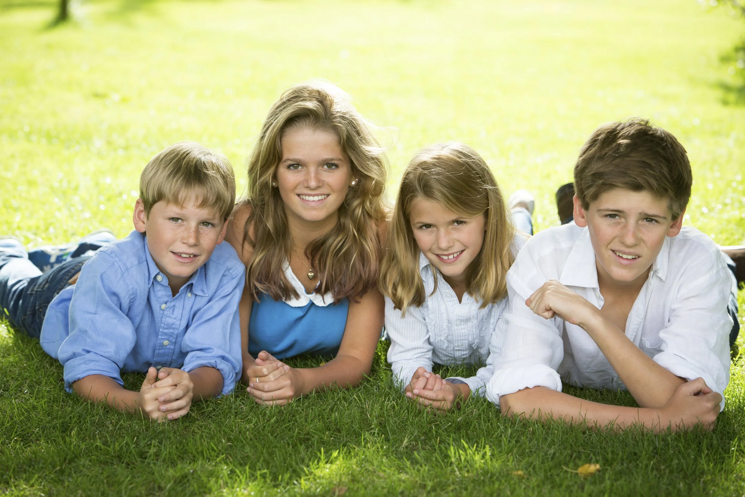 Four children lying on grass outdoors on a sunny day, smiling at the camera.