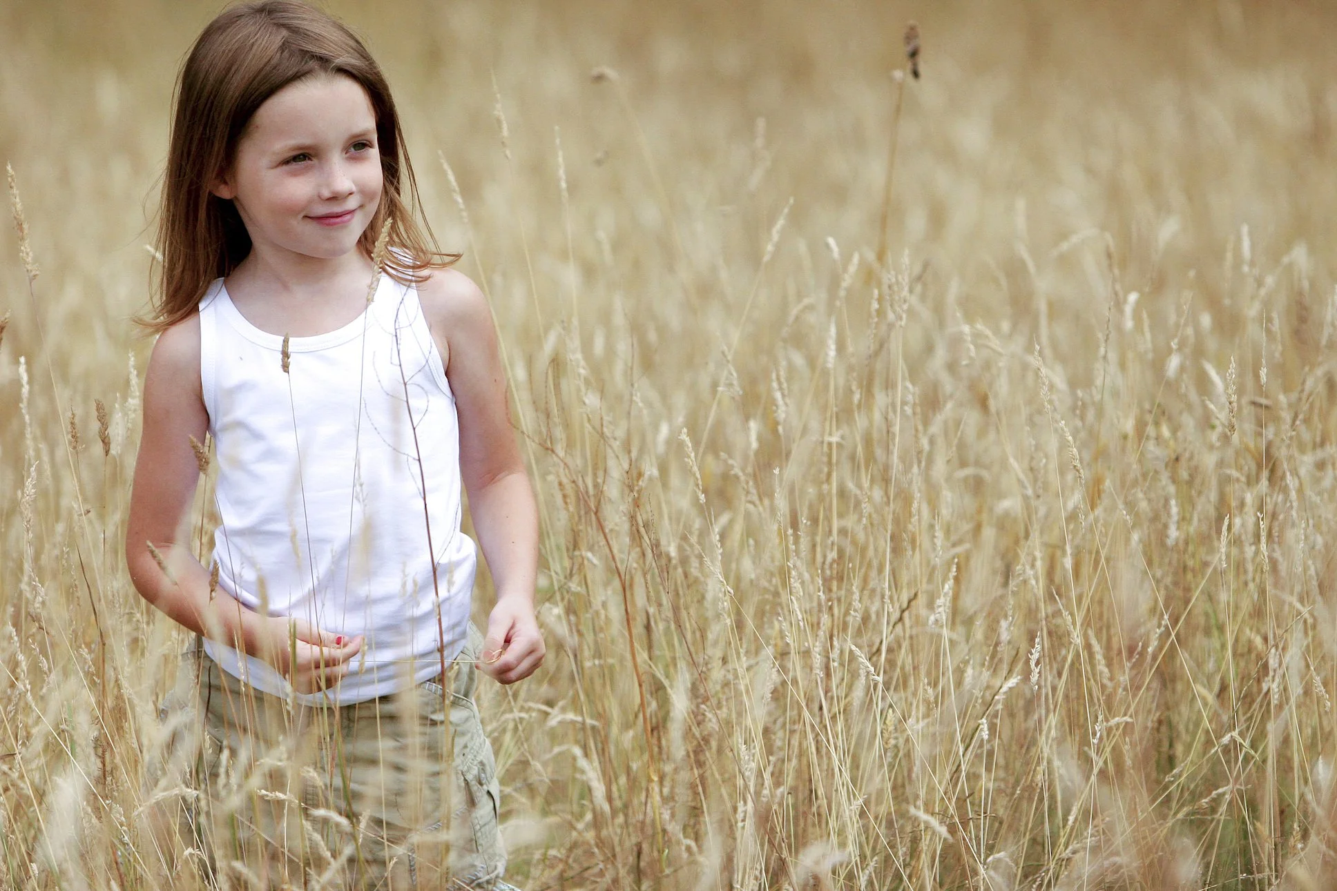 A young girl with light skin and brown hair walking through a field of tall, dry grass.