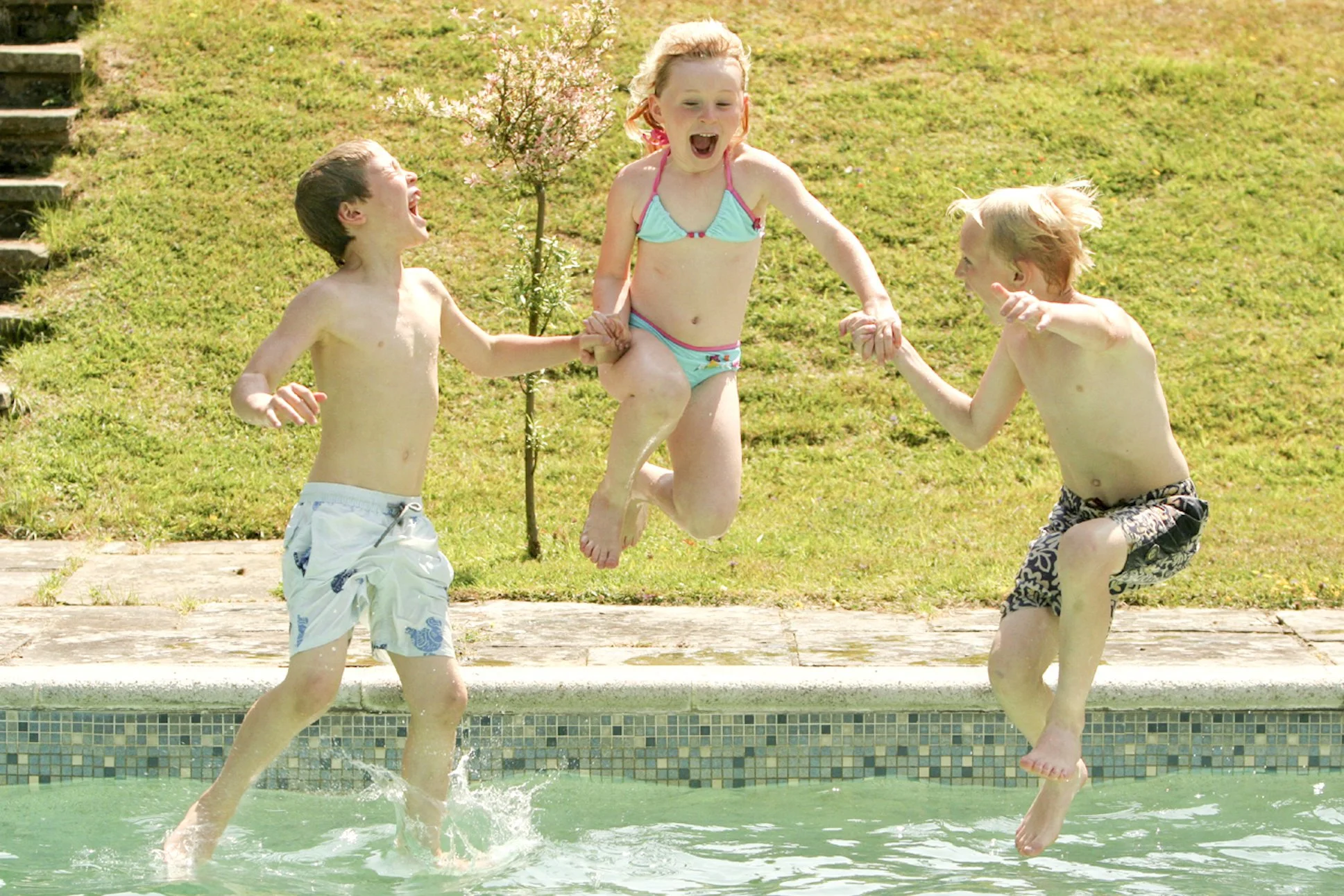 Three children playing and jumping in a swimming pool on a sunny day, holding hands and smiling.