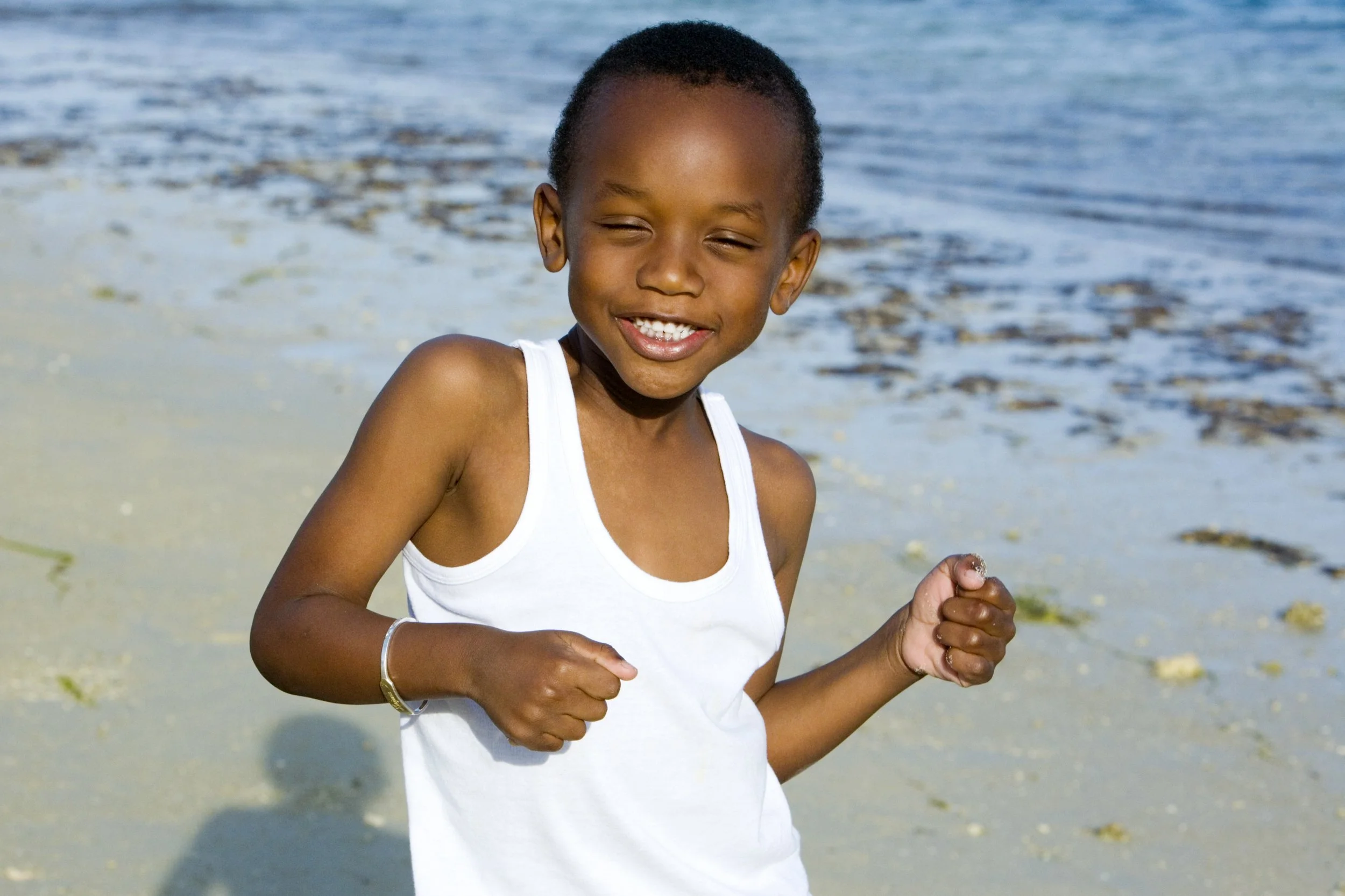 A young boy wearing a white sleeveless shirt, smiling and running on the beach with water and seaweed in the background.