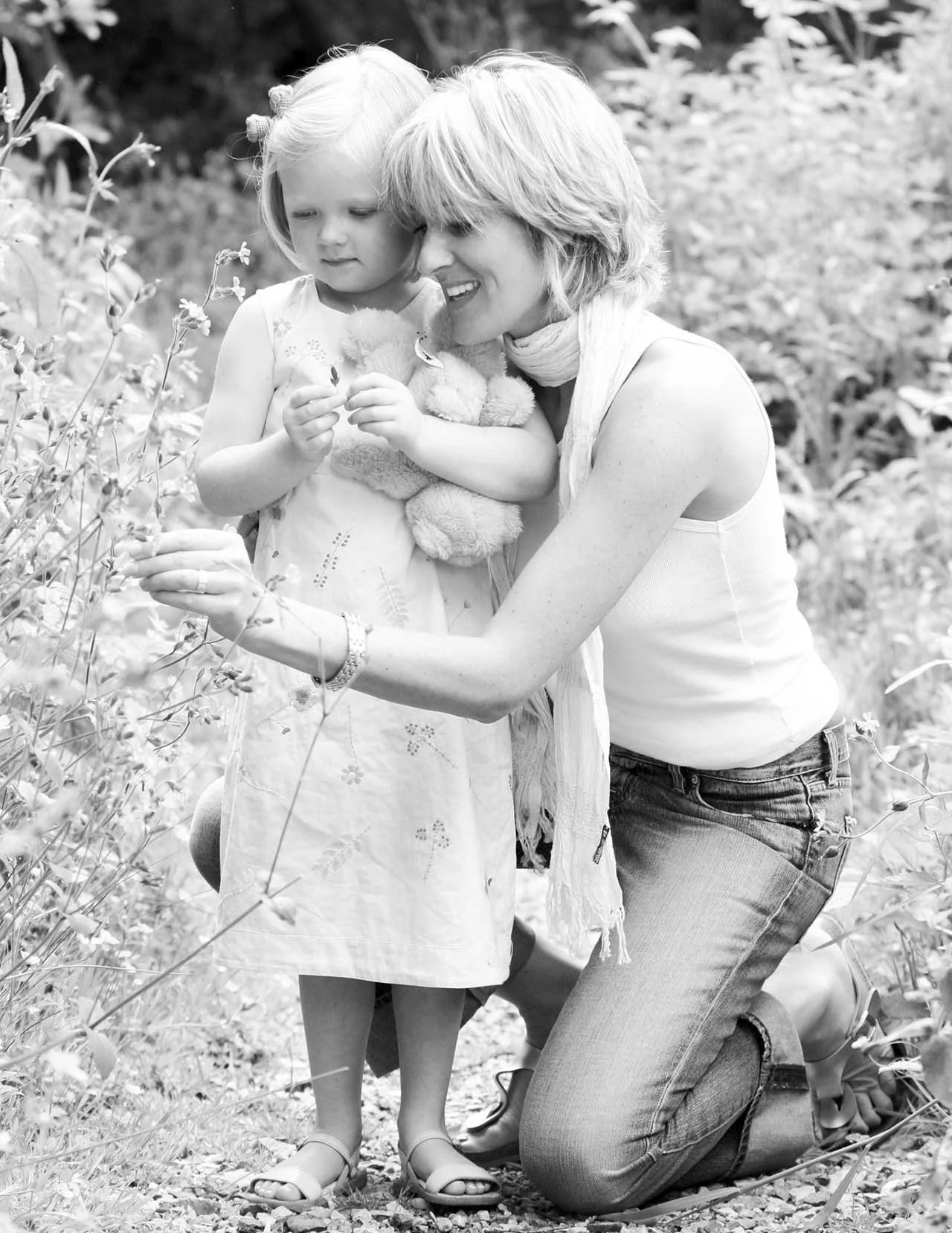 A woman and young girl outdoors among plants, with the woman kneeling and hugging the girl, who is holding a stuffed animal.