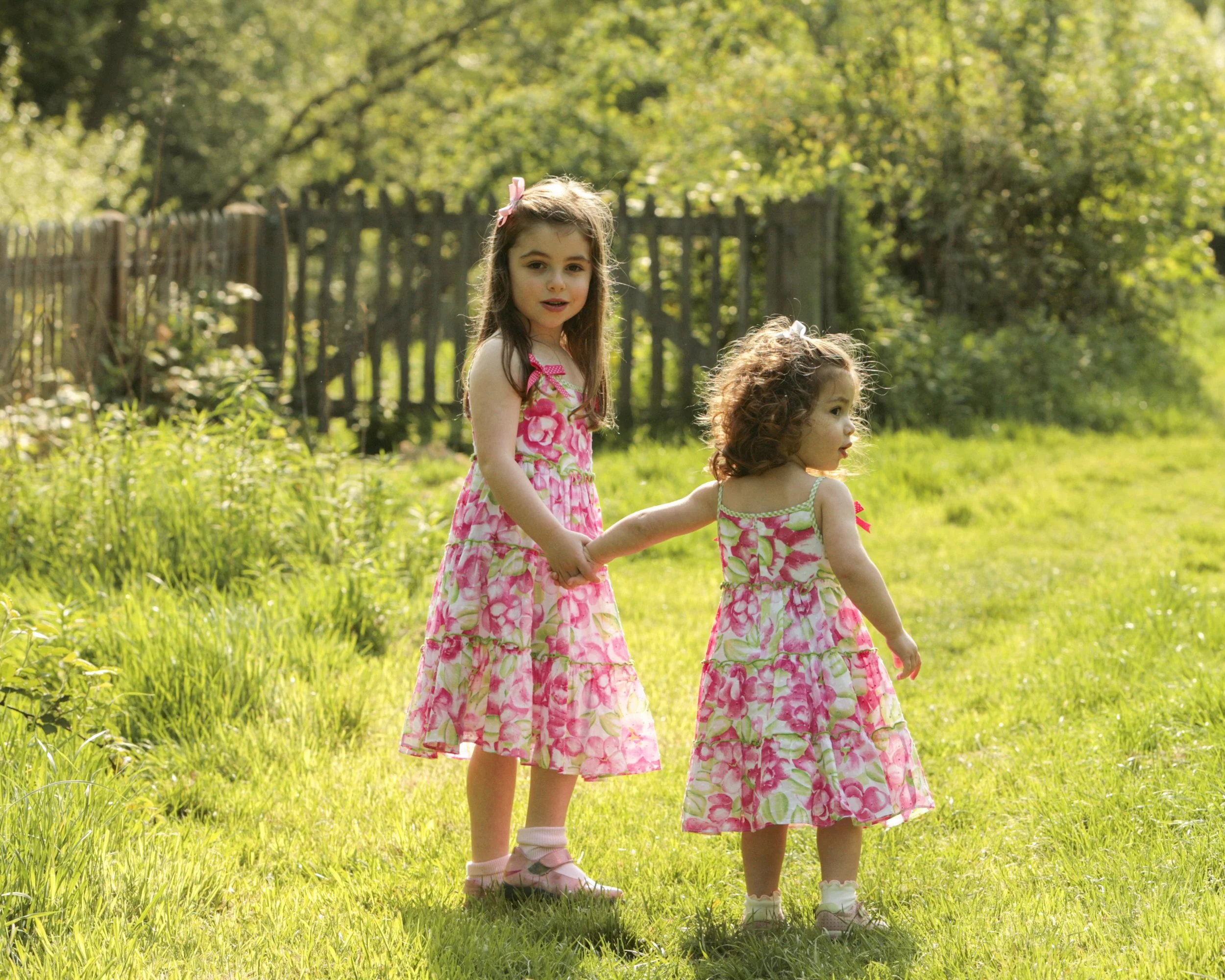 Two young girls in pink floral dresses walking hand in hand on a grassy field in a sunny garden with trees and a wooden fence in the background.