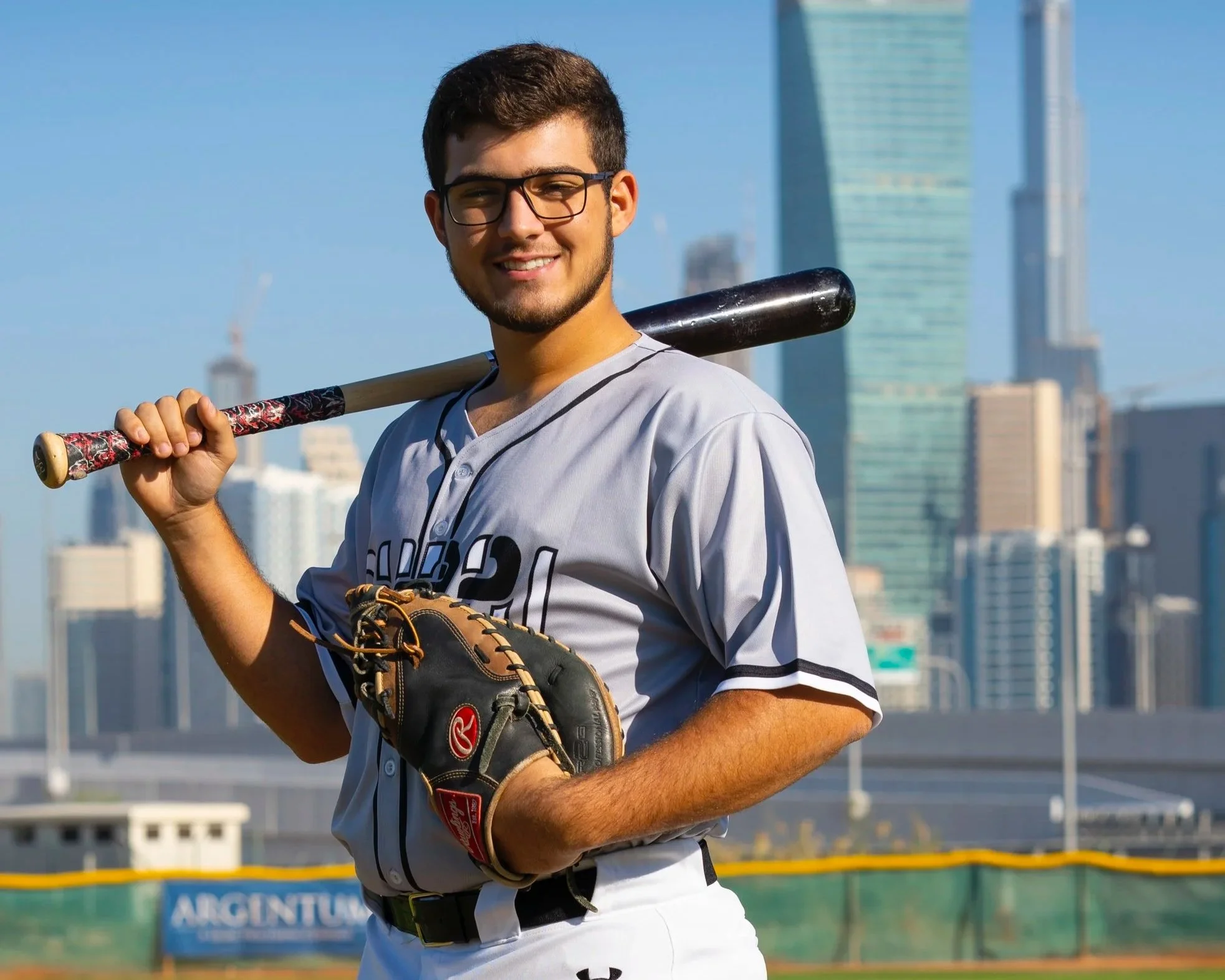 Young male baseball player holding a bat over his shoulder on a city baseball field, wearing glasses, a gray jersey, and a baseball glove on his left hand with a city skyline in the background.