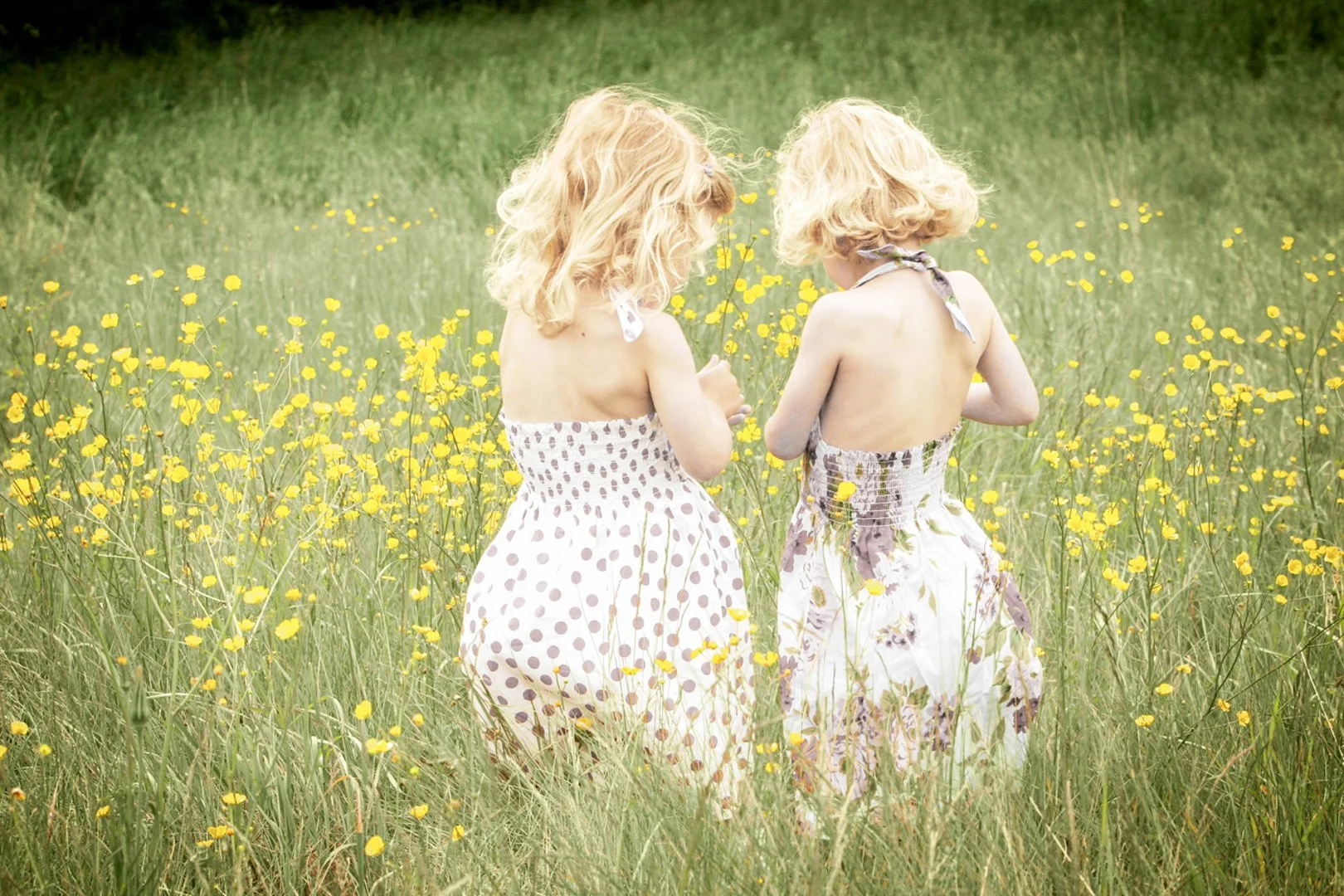 Two young girls with blonde, curly hair walk through a field of yellow flowers, wearing strapless dresses with floral and polka dot patterns.