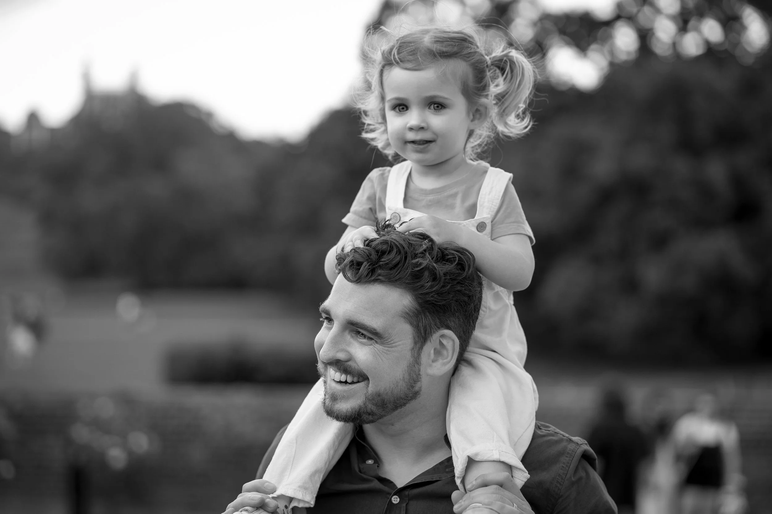 A man carrying a young girl on his shoulders in a park, both smiling, black and white photo.