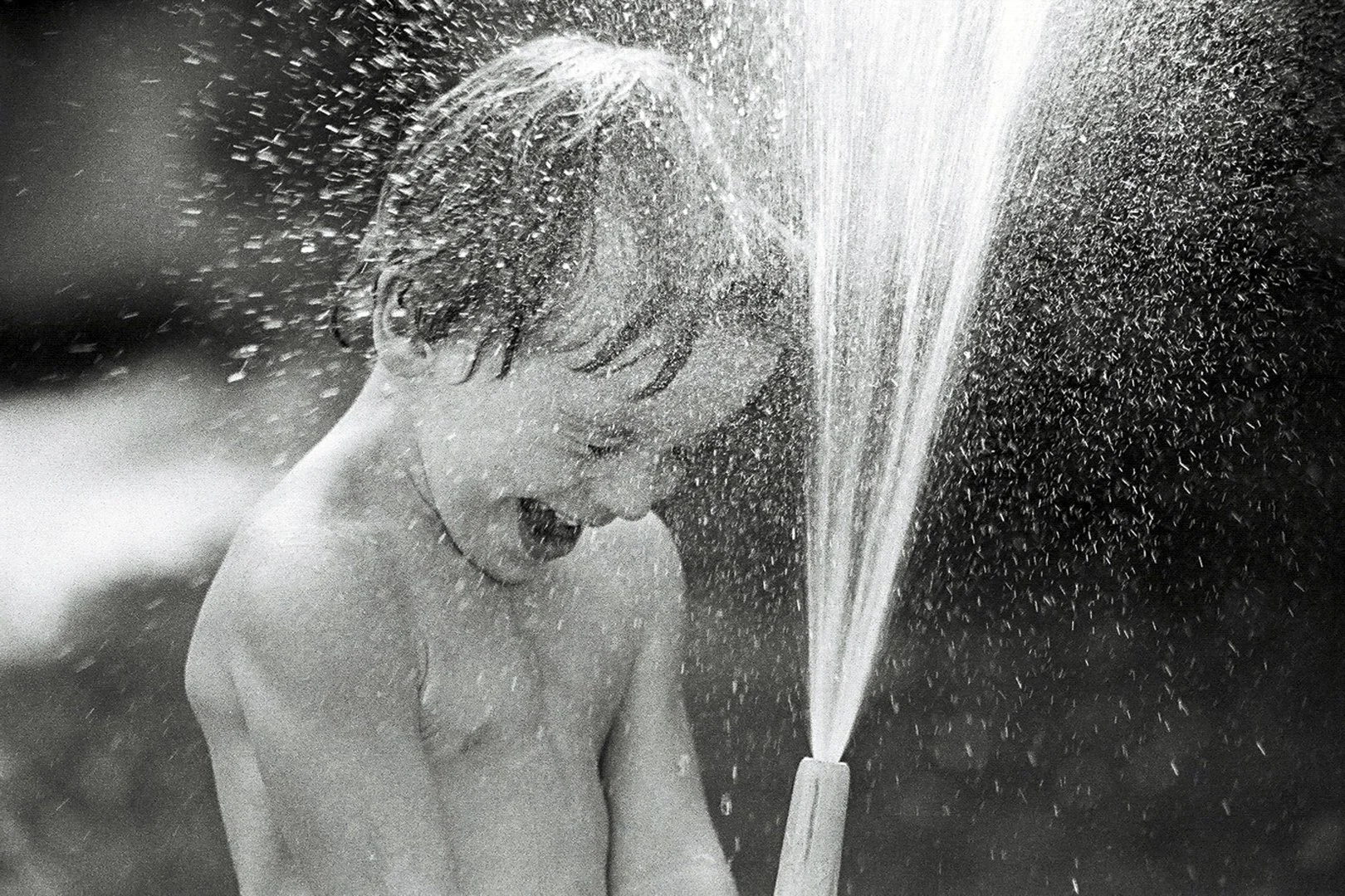 Young boy with wet hair playing with a water spray, closing eyes and smiling.