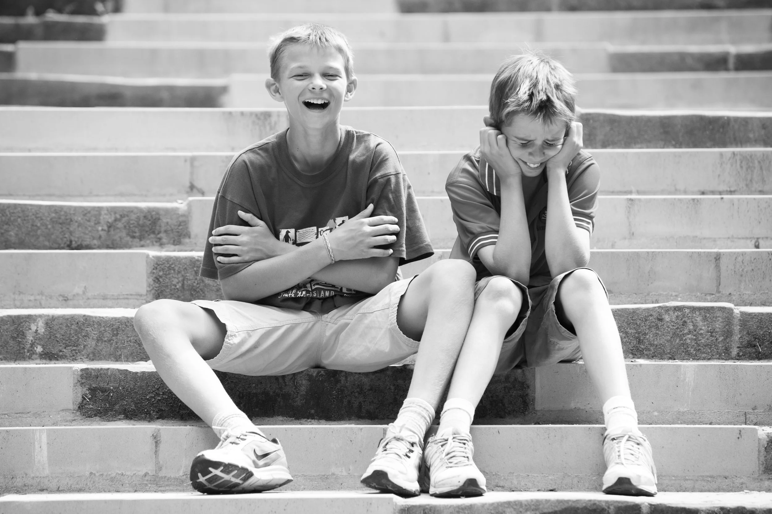 Two boys sitting on outdoor stairs, laughing and covering their ears, enjoying each other's company.