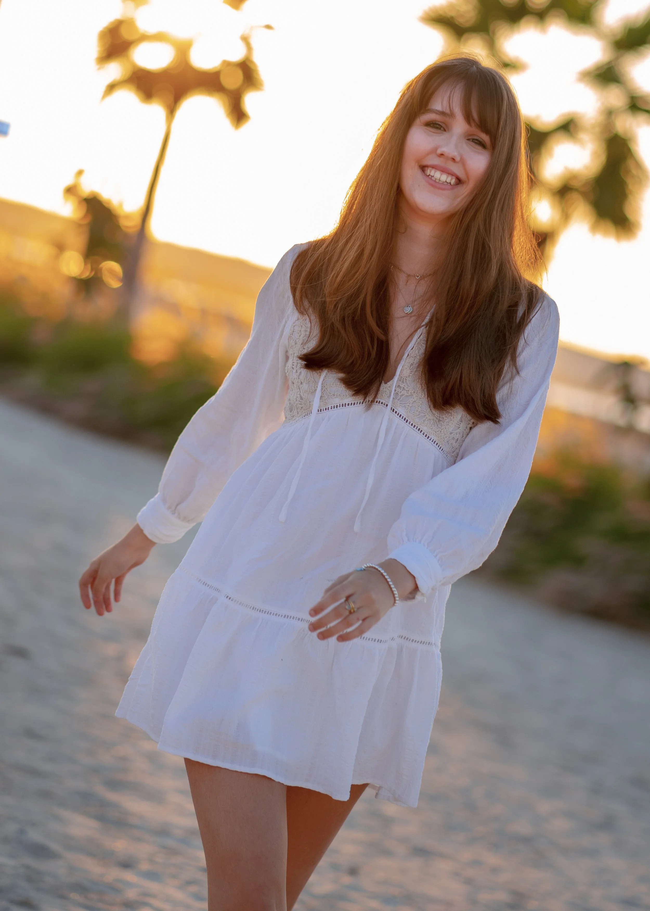 A young woman wearing a white dress smiling and walking outdoors at sunset.