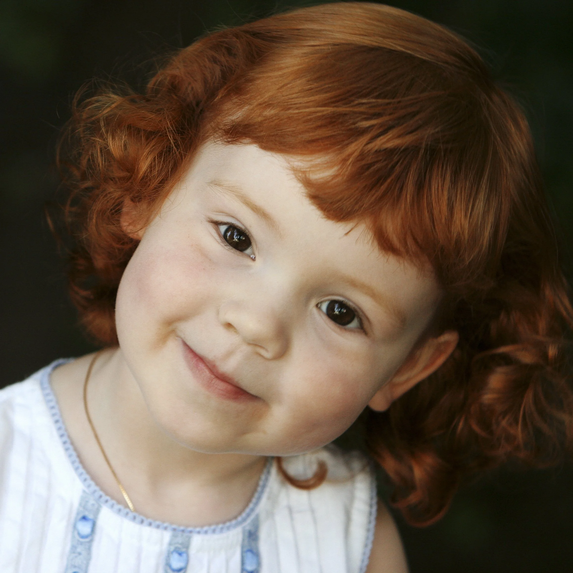 Close-up of a young girl with red hair and a gentle smile, wearing a light-colored dress with blue accents and a gold necklace.