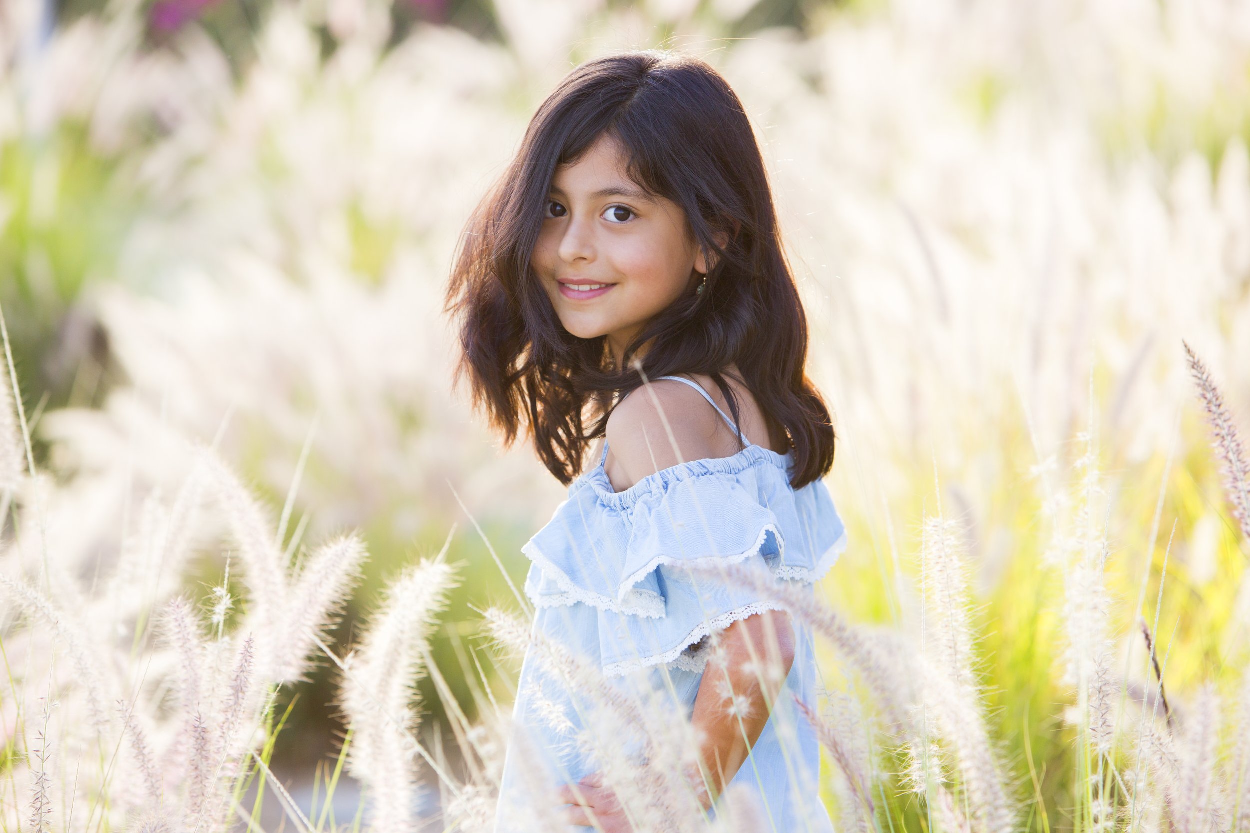 A young girl with dark hair and a light blue off-shoulder dress standing in a grassy field during daylight, smiling at the camera.