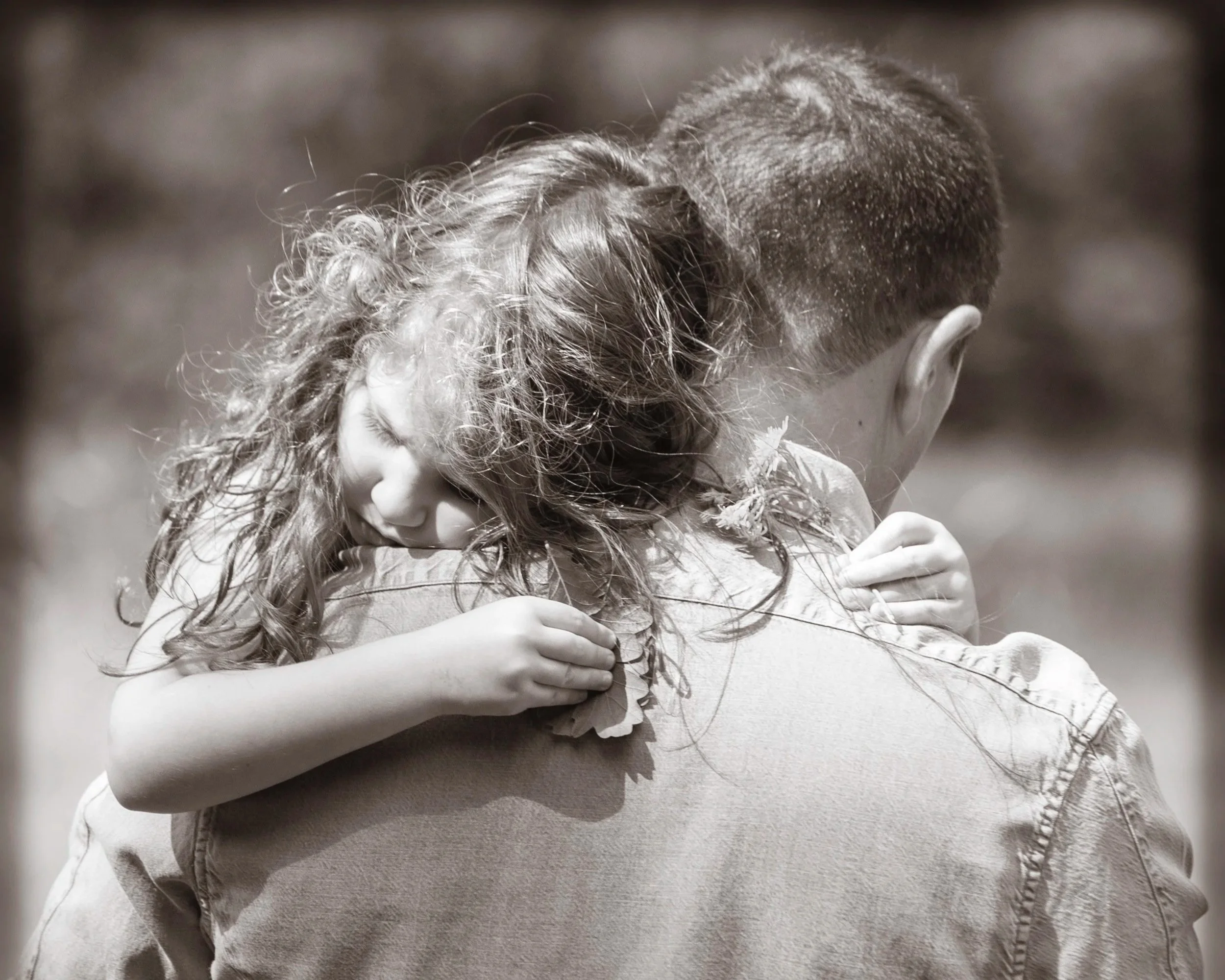 A black and white photo of a young girl hugging a man from behind, resting her head on his shoulder with her eyes closed.